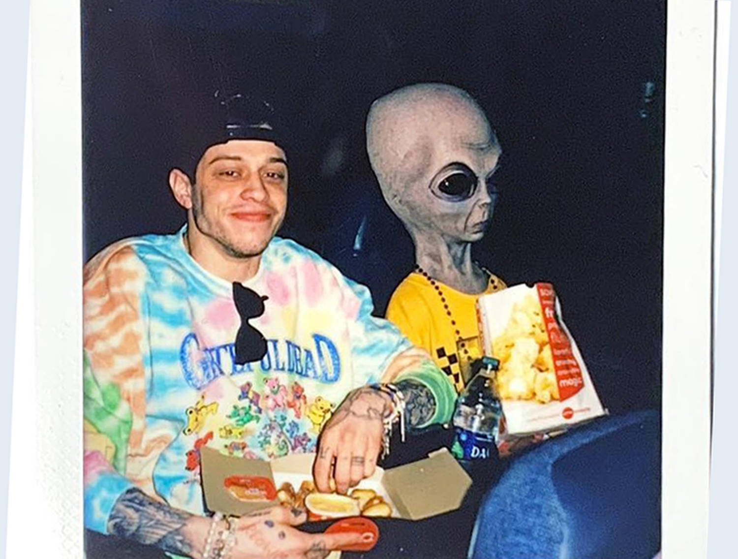 外星人 桌布 壁紙 — 一名穿著紮染襯衫的男子在晚上微笑著吃零食，旁邊是一個穿著外星人服裝的人。