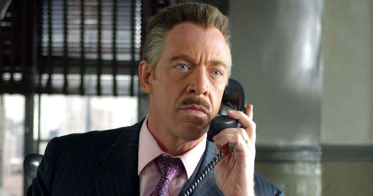 J Jonah Jameson 桌布 壁紙 — 一名穿著細條紋西裝的男子在現代辦公室裡，表情嚴肅地將黑色電話貼在耳邊。