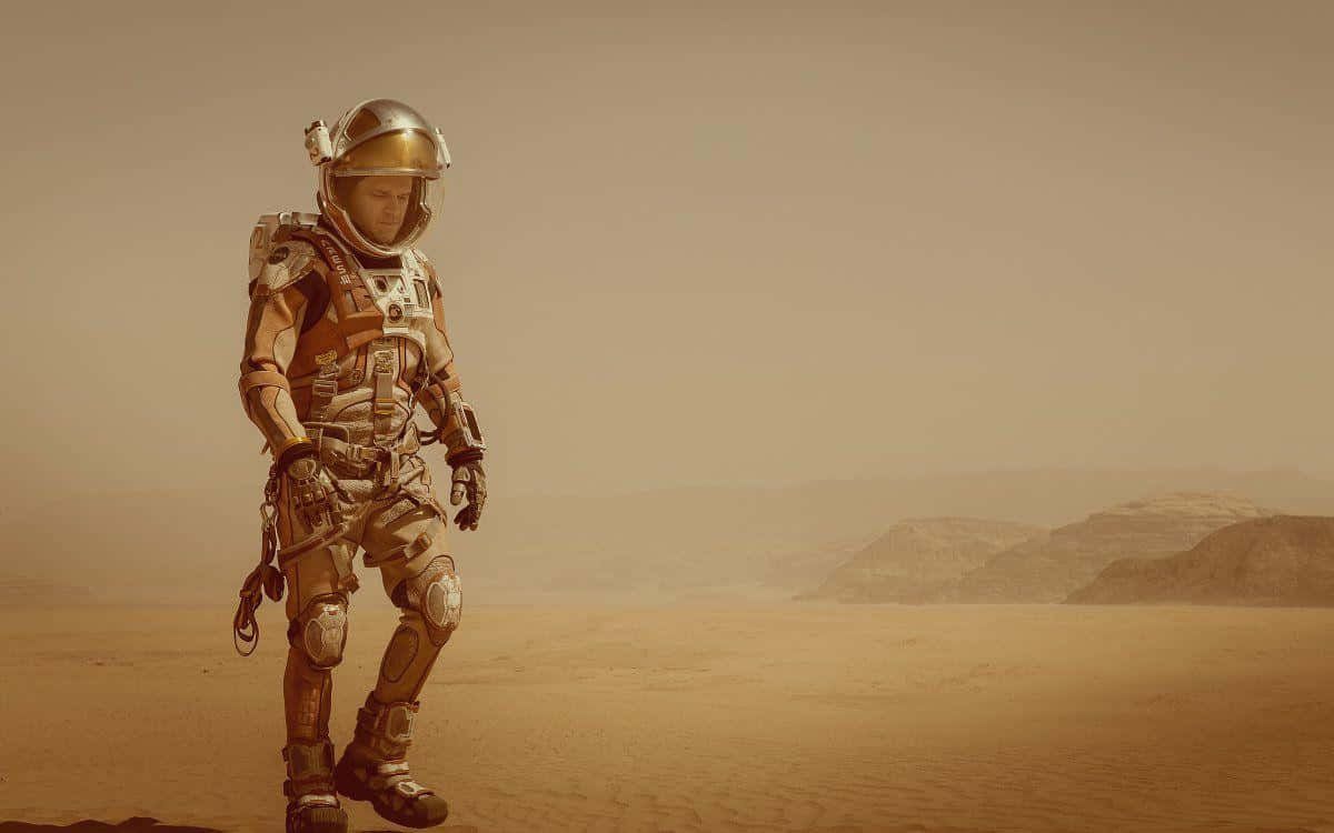 The Martian 桌布 壁紙 — 一名穿著細節豐富太空衣的太空人，在朦朧橙色天空下獨自走過佈滿灰塵的外星沙漠。