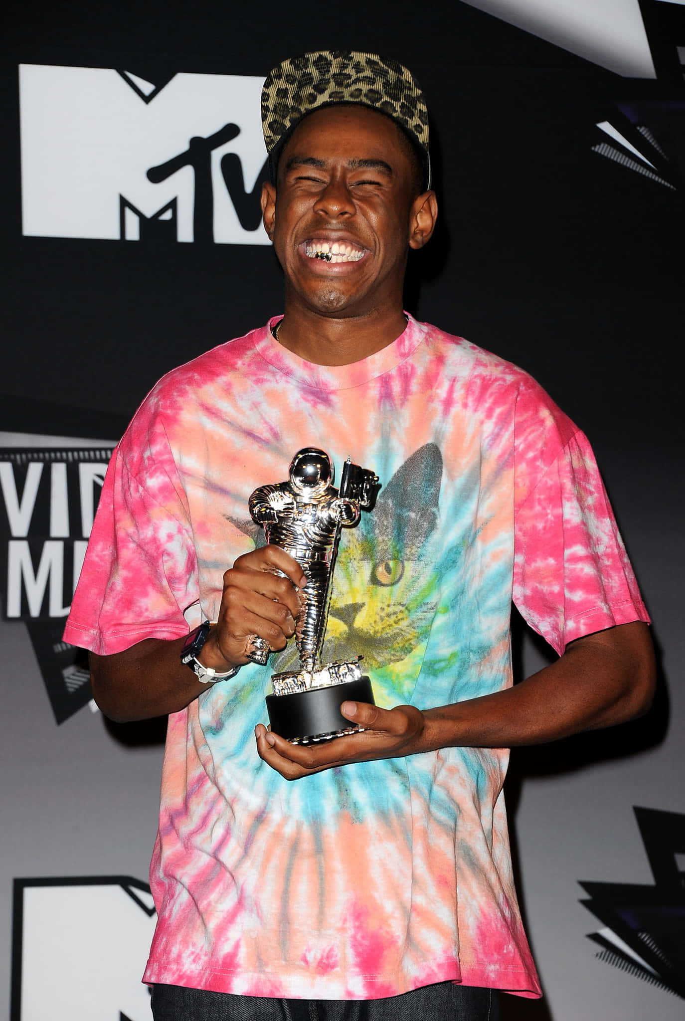 tyler the creator 桌布 壁紙 — 一名穿著鮮豔紮染襯衫的男子在舞台上開懷大笑，手持MTV音樂錄影帶大獎獎盃。