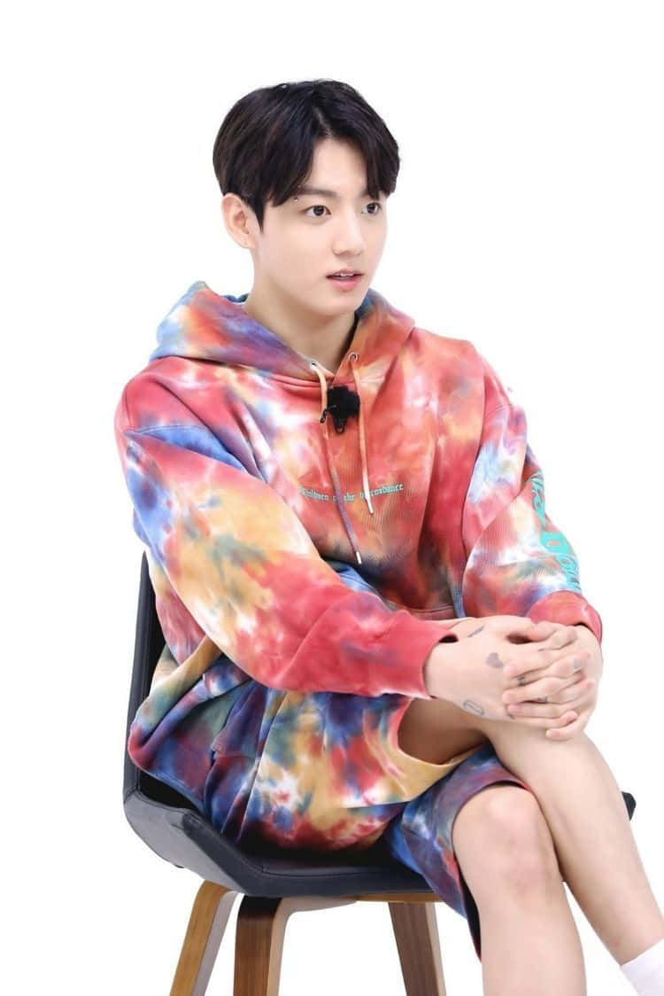 Bts Endorsements 桌布 壁紙 — 一名穿著鮮豔紮染連帽衫的年輕男子，平靜地坐在白色背景前的黑色凳子上。