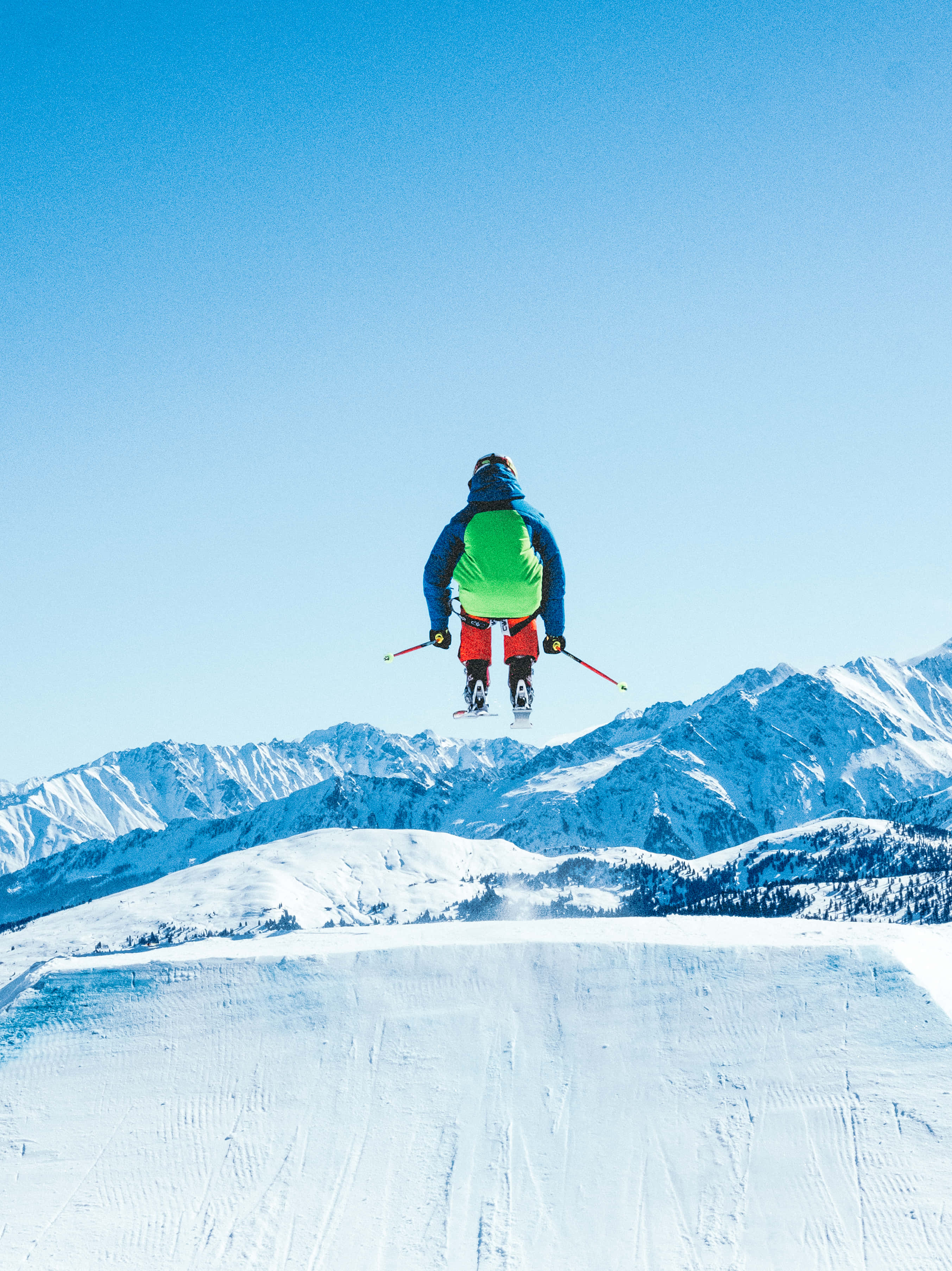 Skiing 桌布 壁紙 — 一名穿著鮮豔裝備的滑雪者在晴朗的天空下，在清澈的藍色空氣中飛越白雪覆蓋的山脈。