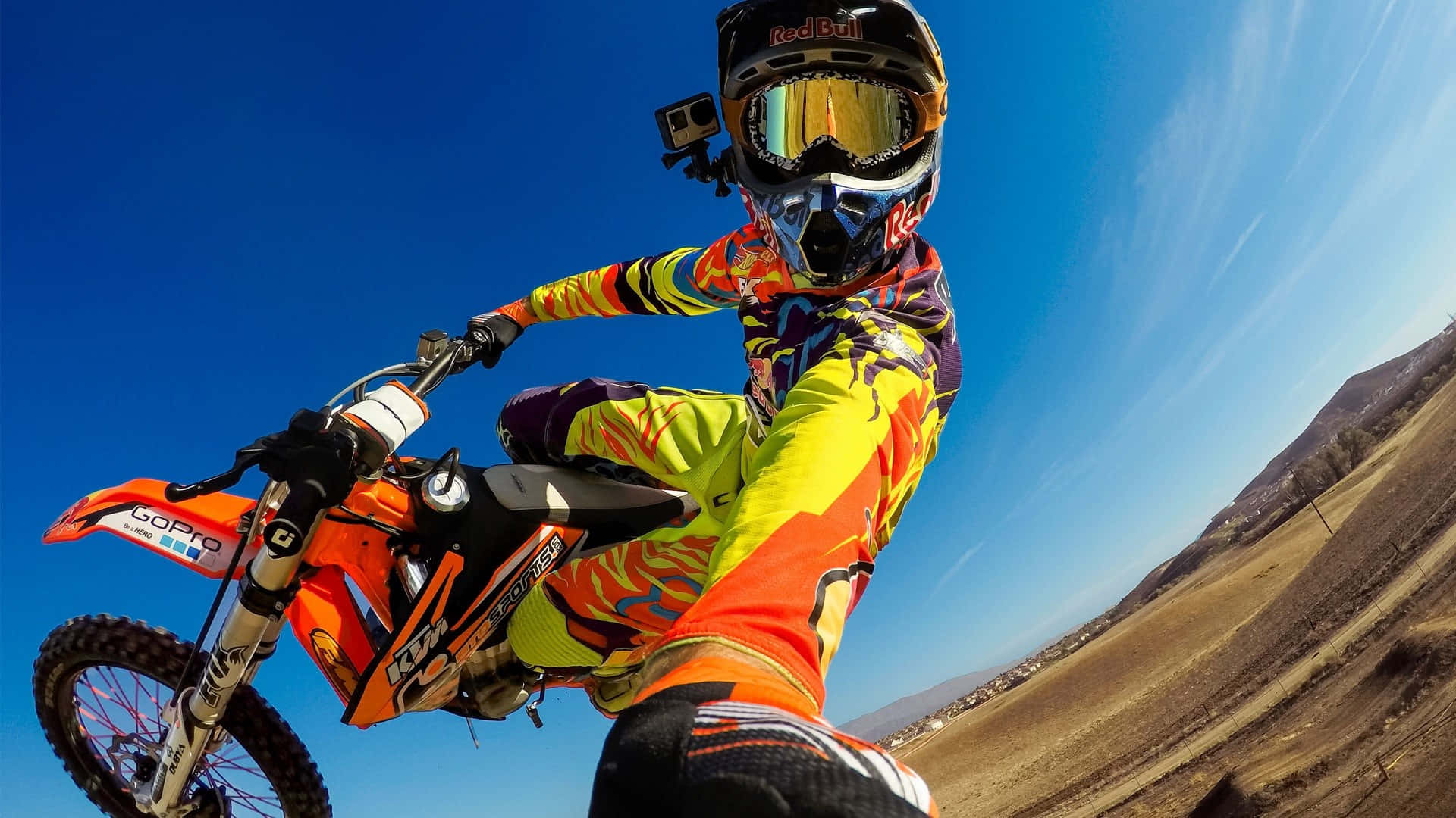 Motocross 桌布 壁紙 — 一名穿著鮮豔裝備的越野電單車手在橙色越野車上傾斜入彎，背景是清澈的藍天。