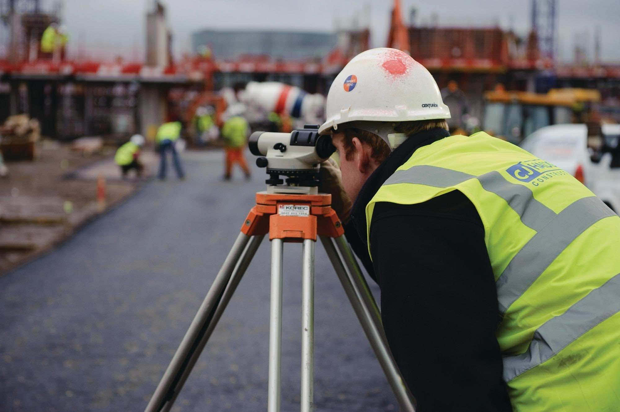 Engineer 桌布 壁紙 — 一名穿著黃色背心及安全帽的建築工人在繁忙的建築工地使用三腳架上的測量望遠鏡。