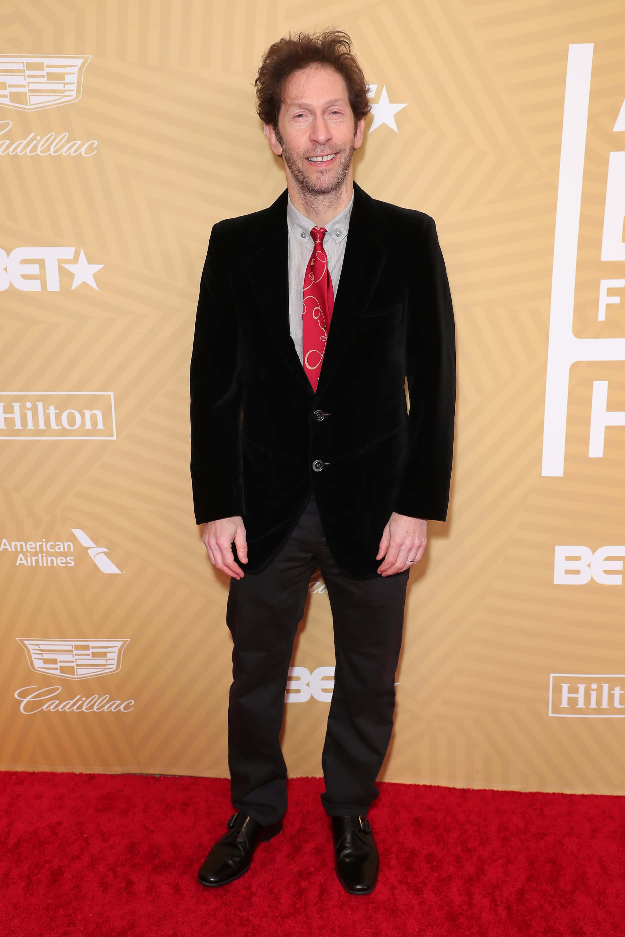 Tim Blake Nelson 桌布 壁紙 — 一名穿著黑色天鵝絨西裝和紅色領帶的男子在品牌金色背景前的紅地毯上微笑。