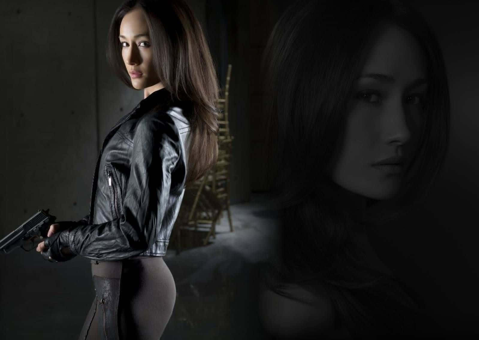 maggie q 桌布 壁紙 — 一名穿著黑色皮褸的女子在光線昏暗的工業空間中手持槍支，並帶有鬼臉疊加效果。