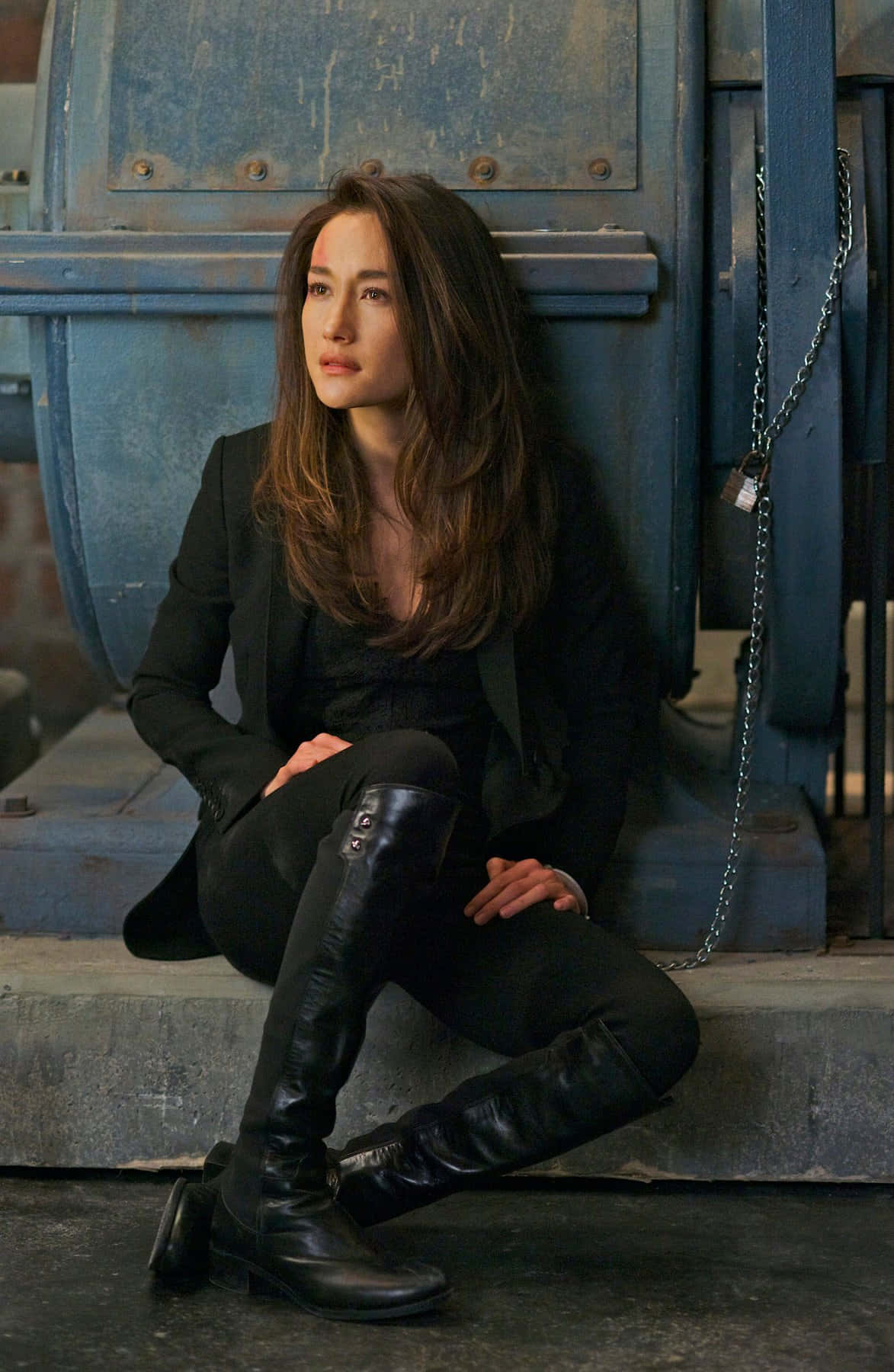 maggie q 桌布 壁紙 — 一名穿著黑色皮靴和西裝的女性，若有所思地靠在一扇帶有鏈鎖的風化藍色工業門上。