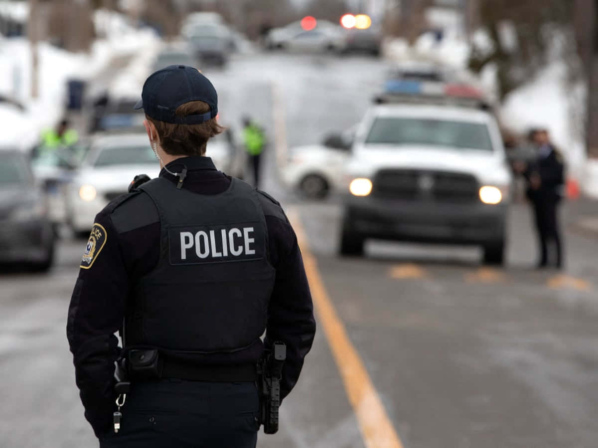警察 桌布 壁紙 — 一名穿著黑色背心的警察站在積雪的街道上，一輛閃著警示燈的巡邏車正在靠近。