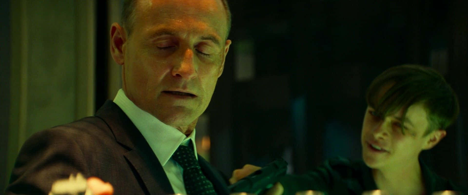 Harry Osborn 桌布 壁紙 — 一名穿西裝的男子閉著眼睛拿著槍，而一名微笑的年輕男子站在他身後，房間內有昏暗的綠色燈光。