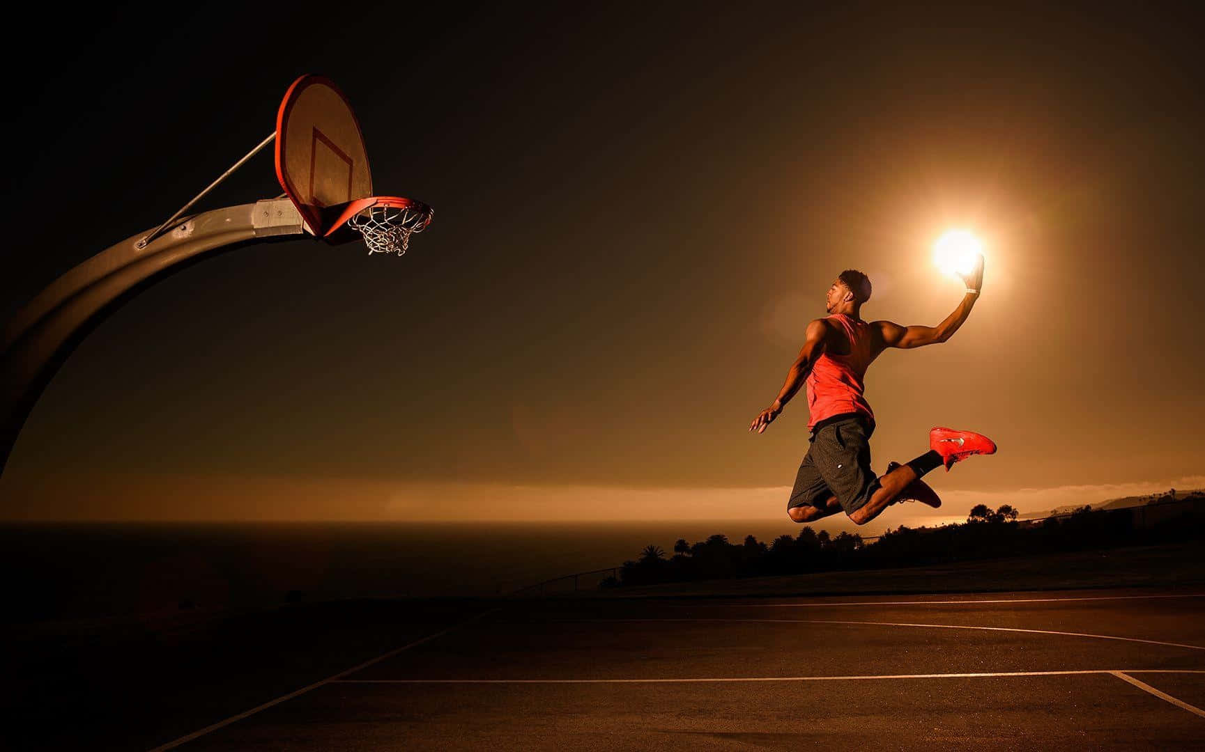 Cool Basketball 桌布 壁紙 — 一名籃球運動員在夜晚的戶外球場上，在街燈下拿著發光的球跳向籃框。