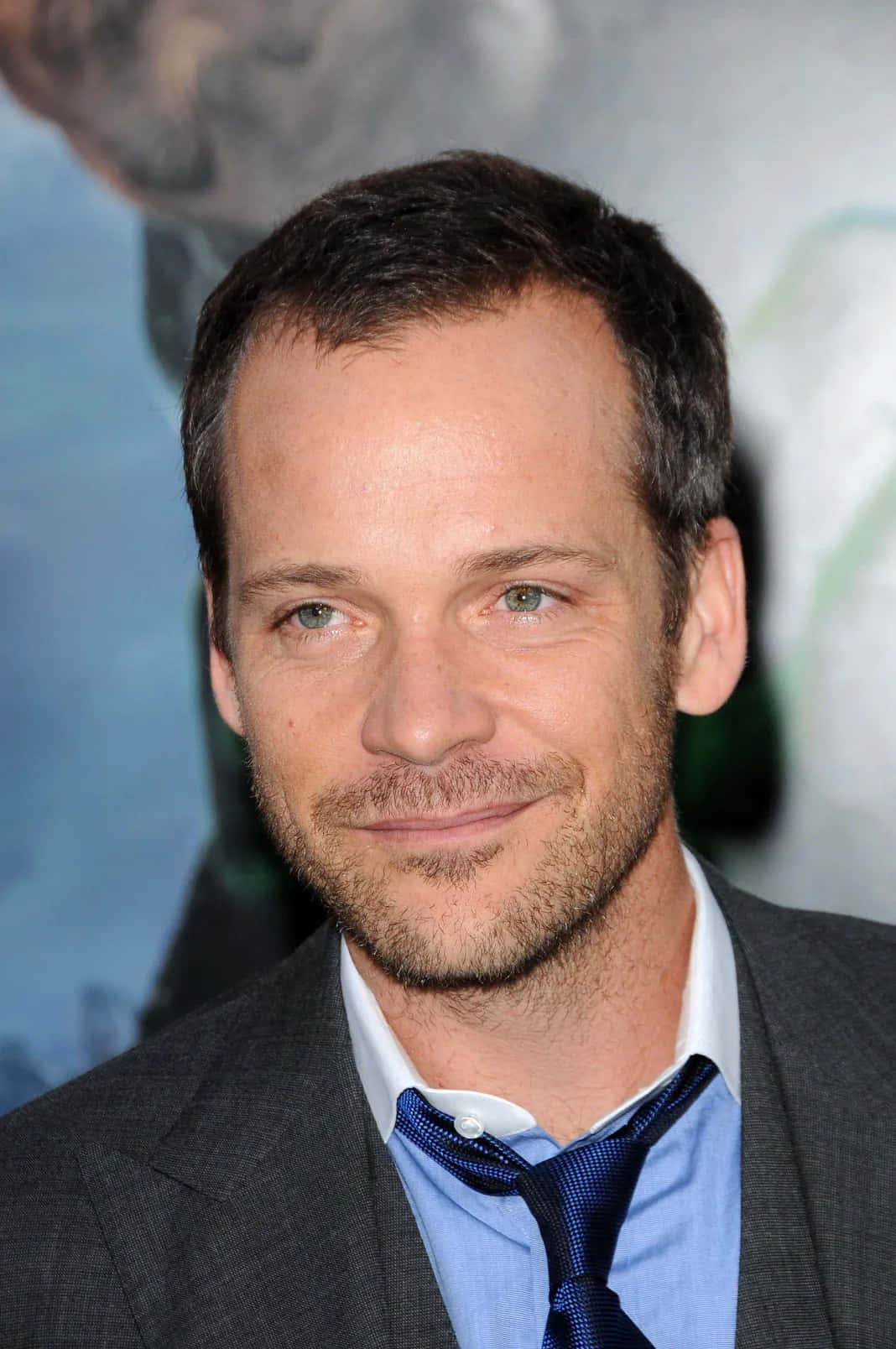 Peter Sarsgaard 桌布 壁紙 — 一名綠眼鬍渣男士的特寫肖像，身穿灰色西裝、藍色襯衫和藍色領帶。