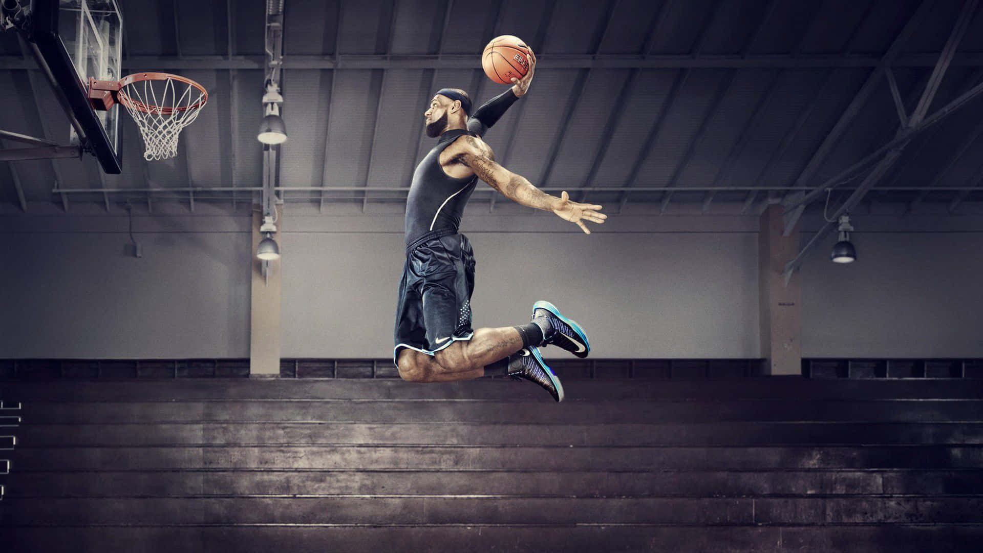 Best Sports 桌布 壁紙 — 一名肌肉發達的籃球運動員在室內球場高高躍起，準備在工業照明下，於空無一人的看台上方灌籃。