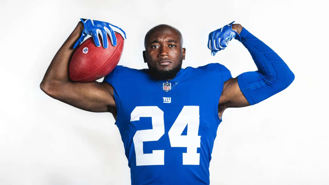 James Bradberry 桌布 壁紙 — 一名肌肉發達的足球員身穿藍色紐約巨人隊球衣，手持紅色足球，在純白背景前展示肌肉。