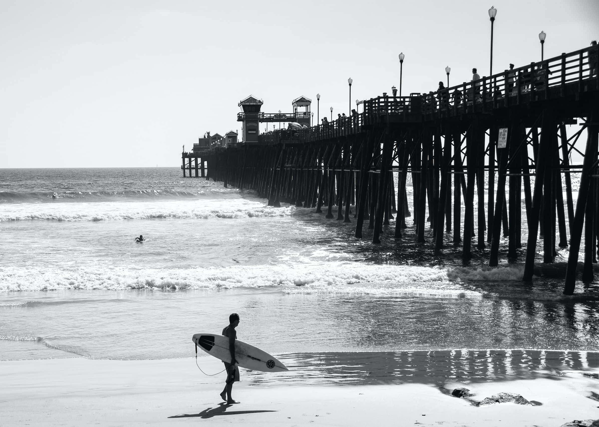 Black And White Beach 桌布 壁紙 — 一名衝浪者喺陽光普照嘅沙灘上行走，帶住一塊衝浪板，海浪喺一條長木碼頭附近拍打。