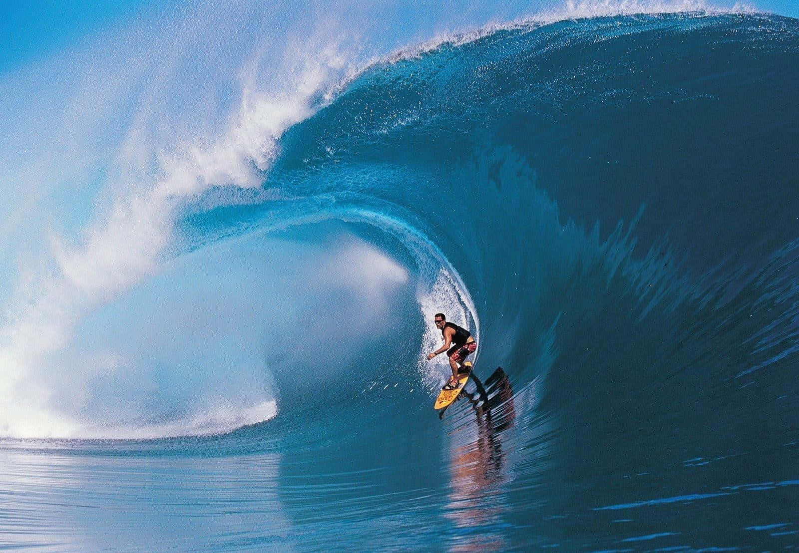 surfing 桌布 壁紙 — 一名衝浪者在蔚藍的天空下，駕馭著巨大的藍綠色海浪的浪管，水花四濺。