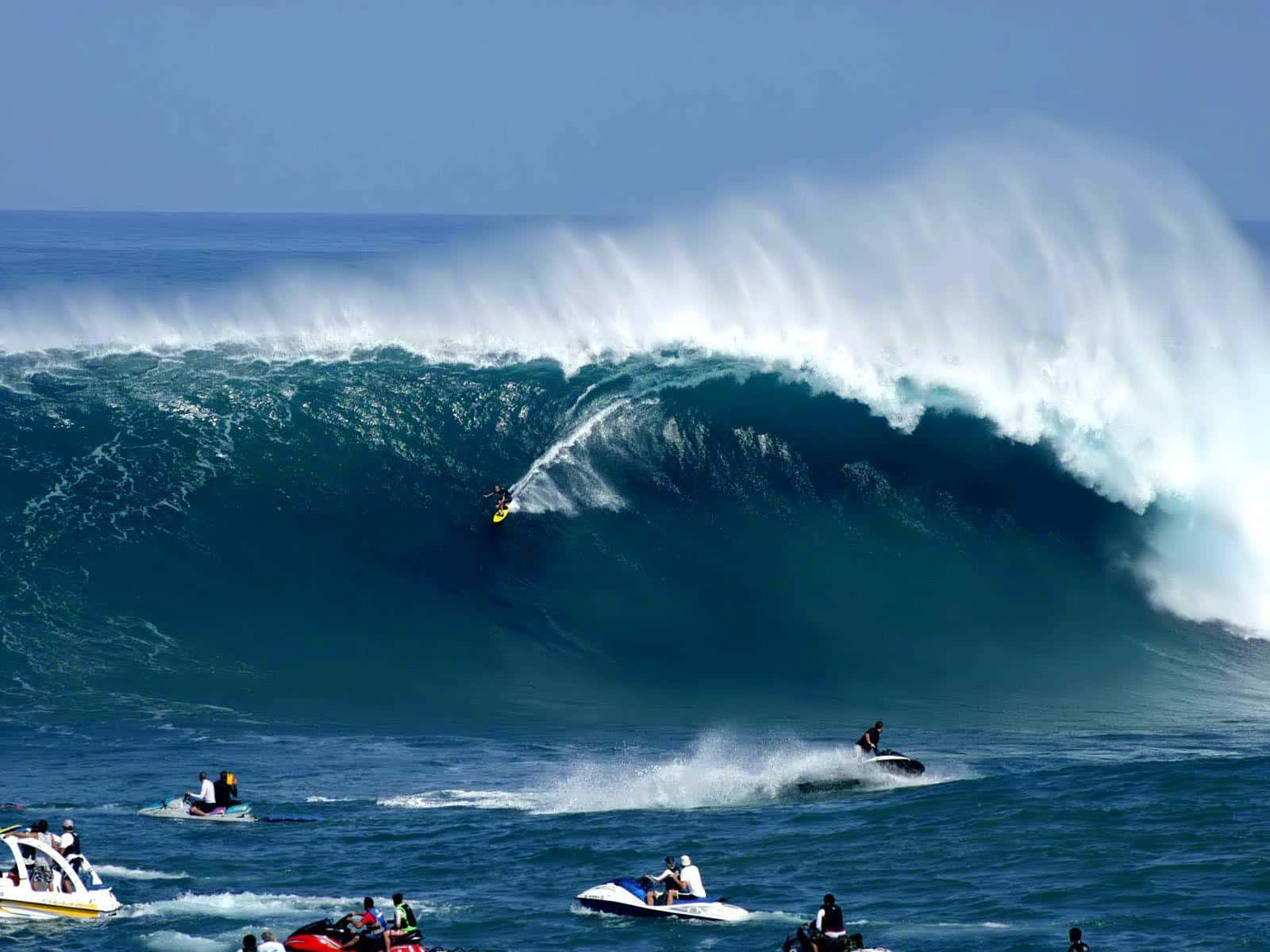 surfing 桌布 壁紙 — 一名衝浪者騎乘巨大的藍綠色海浪，水上摩托車和船隻在下方觀看，天空清澈湛藍。
