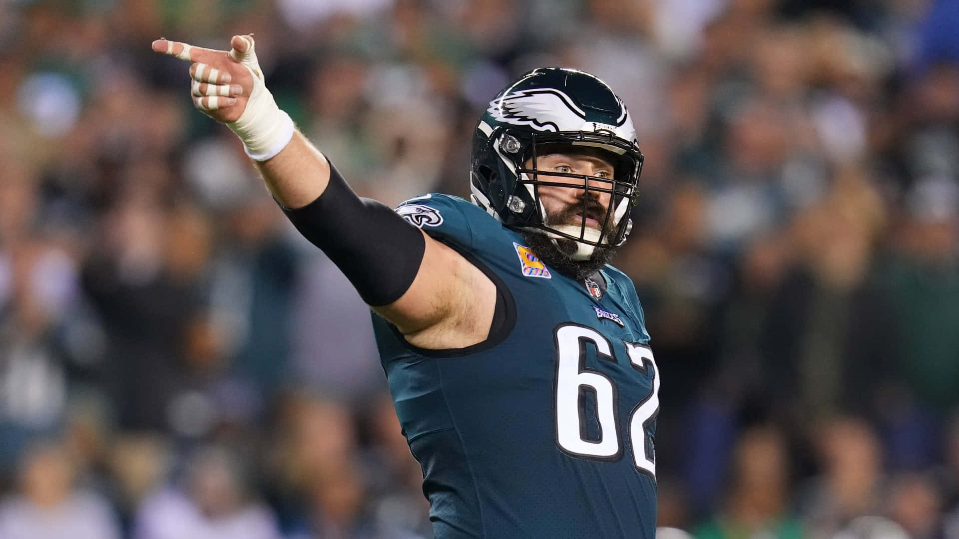 Jason Kelce 桌布 壁紙 — 一名費城老鷹隊美式足球員身穿青綠色62號球衣，在球場燈光下比賽時堅定地向上指。
