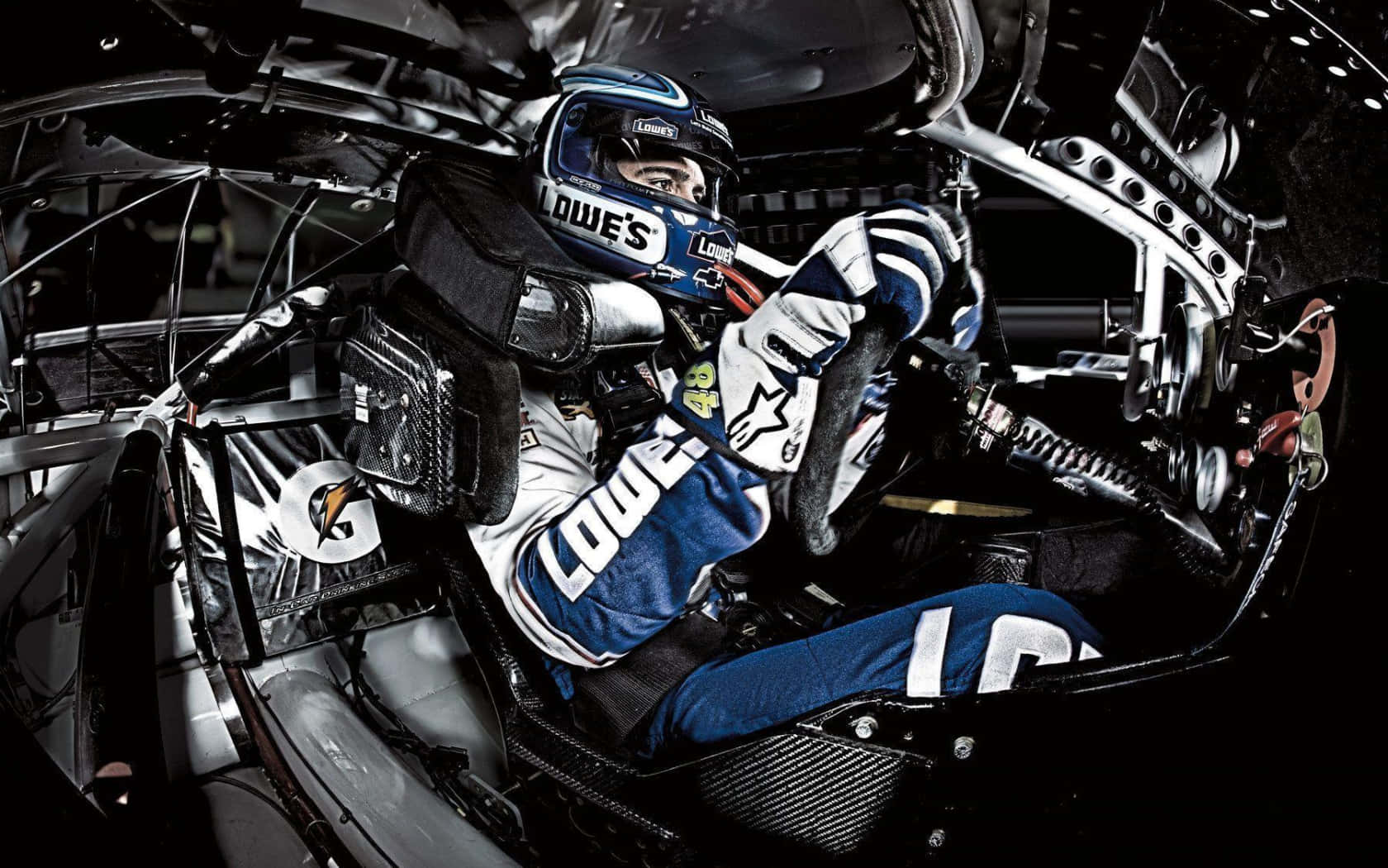 Jimmie Johnson 桌布 壁紙 — 一名賽車手戴著Lowe's品牌的頭盔和賽車服，在細節豐富的量產車駕駛艙內緊握方向盤。