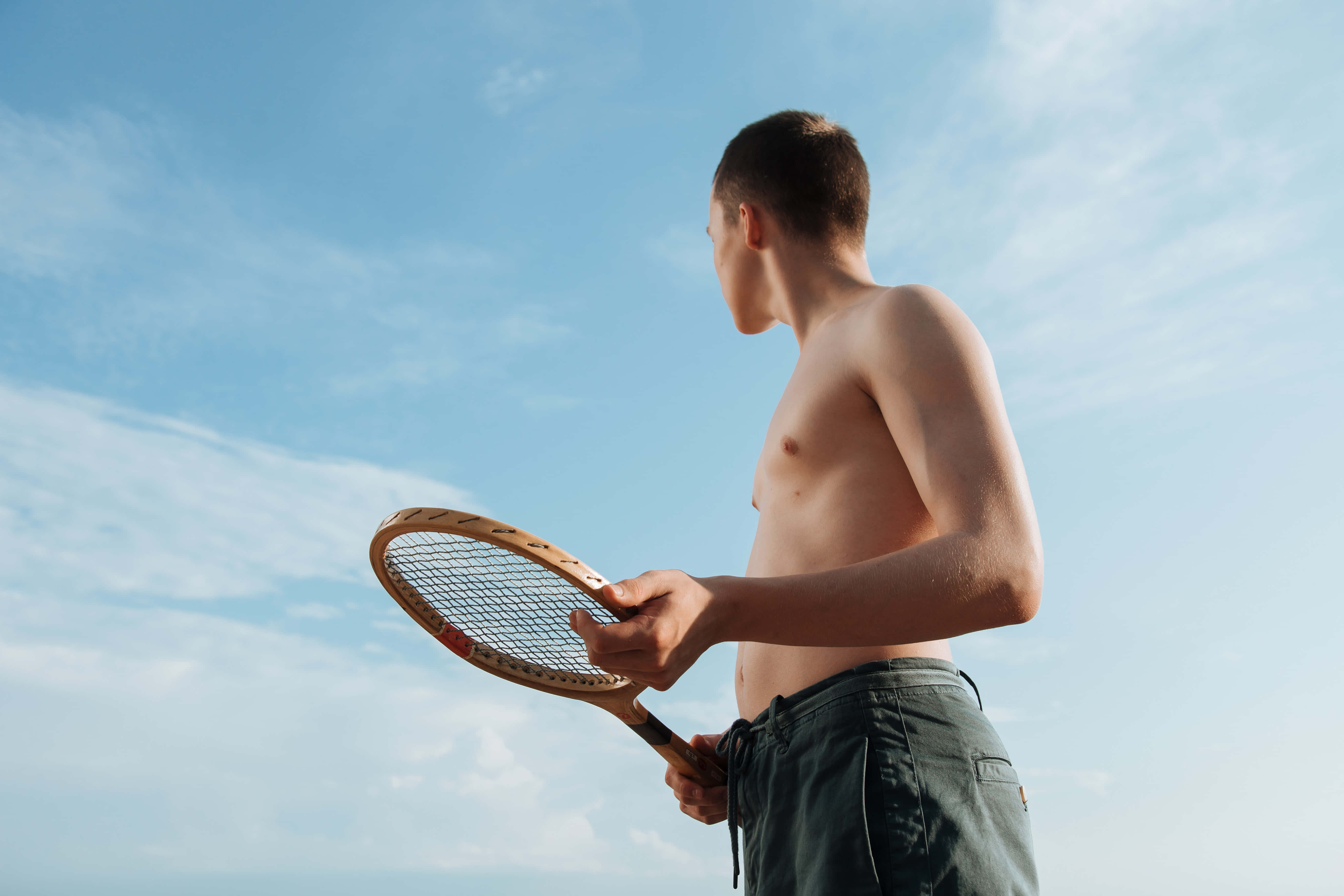 Badminton 桌布 壁紙 — 一名赤裸上身的年輕男子手持木製網球拍，背景是帶有縷縷白雲的明亮藍天。