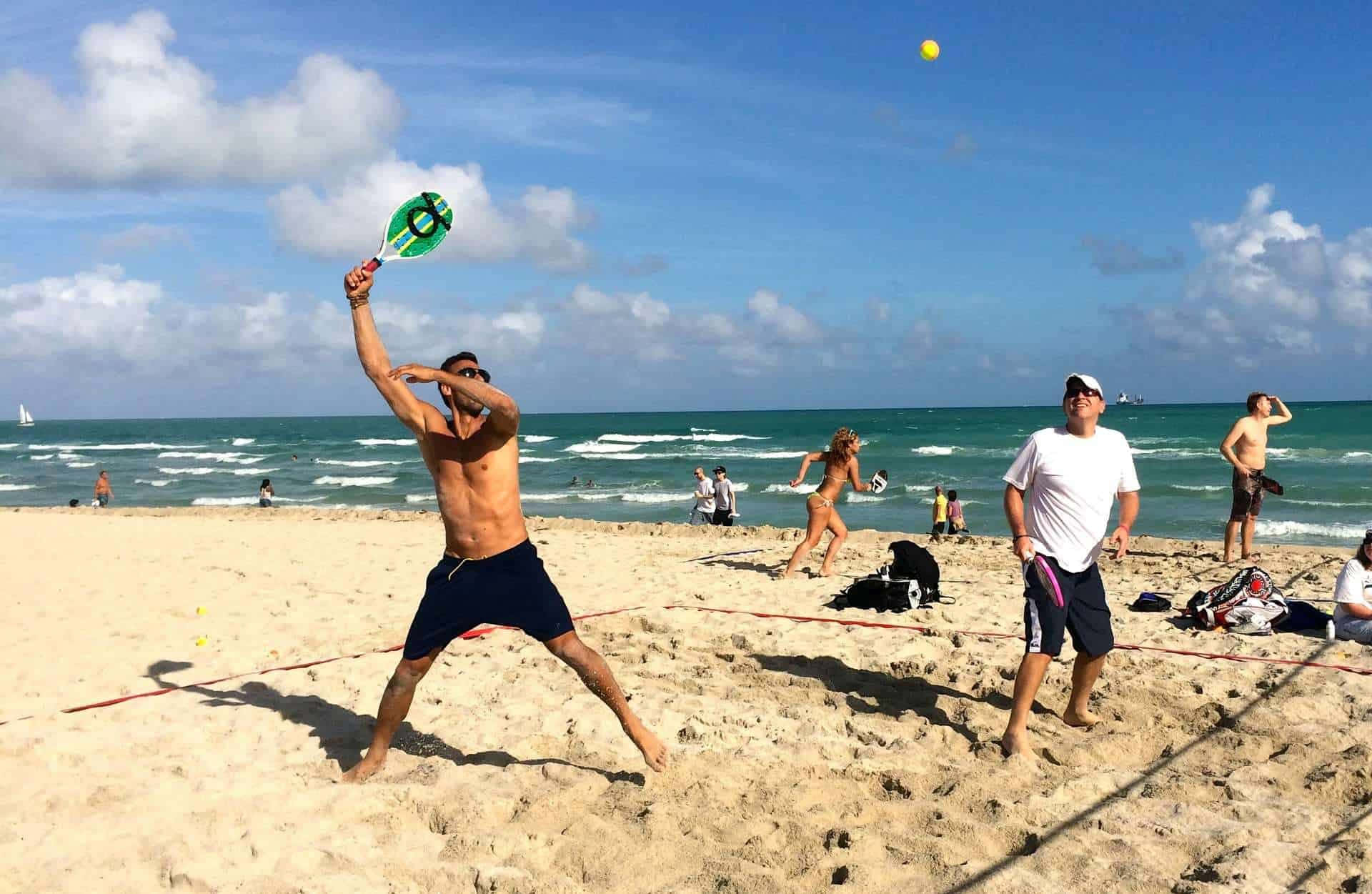Beach Tennis 桌布 壁紙 — 一名赤裸上身的男子在明亮的藍天下，於一場熱鬧的沙灘排球比賽中揮動綠色球拍擊打黃色球。