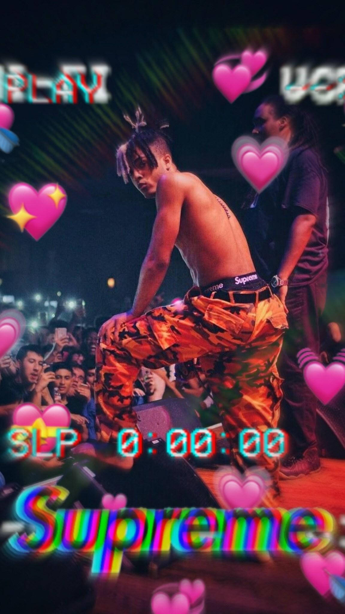 XXXTENTACION 桌布 壁紙 — 一名赤裸上身的男子穿著橙色迷彩褲，在霓虹燈光下，伴隨著漂浮的粉紅色心形和「Supreme」標誌，在舞台上充滿活力地跳舞。