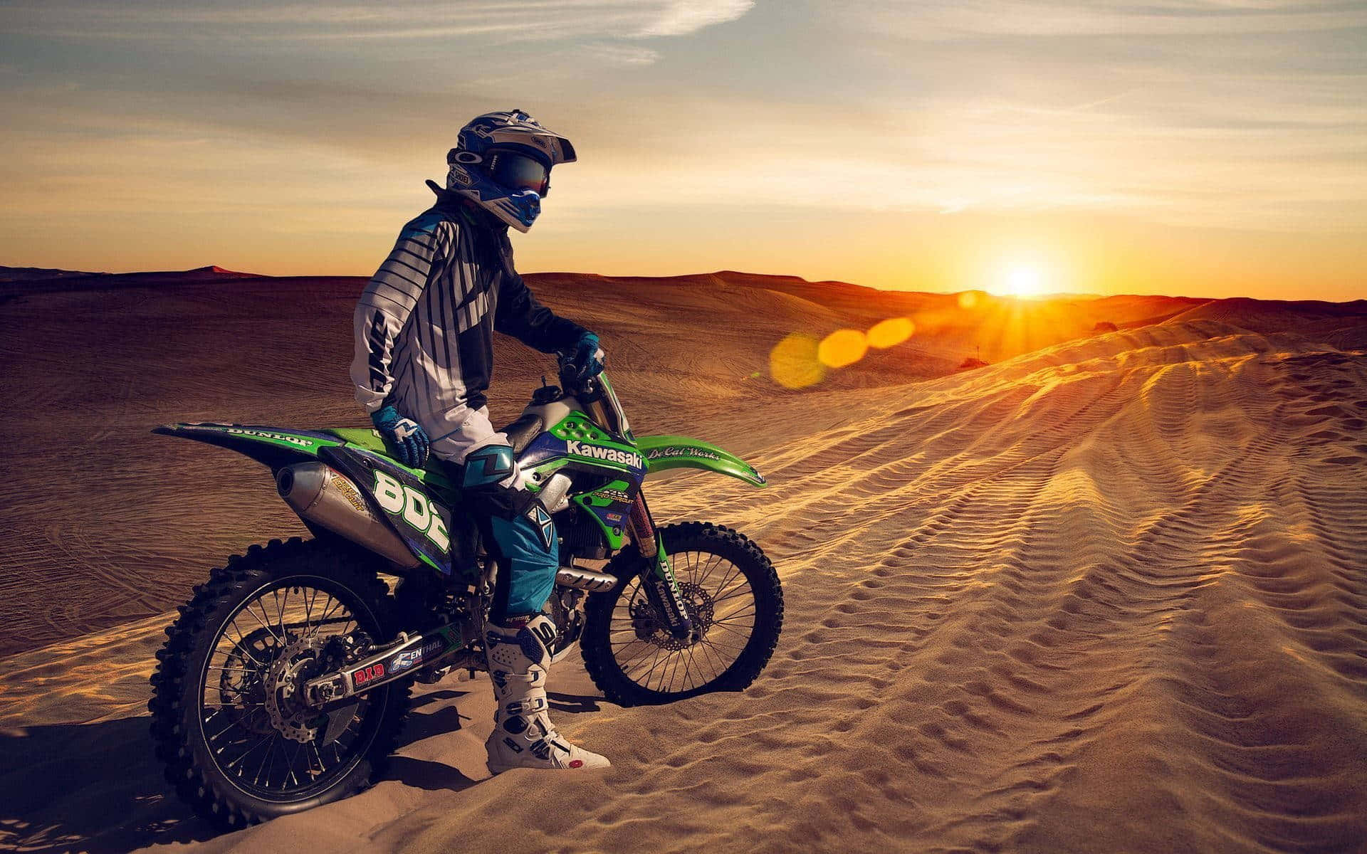 Motocross 桌布 壁紙 — 一名越野電單車手騎著綠色川崎電單車，在沙丘頂上稍作停留，夕陽在他身後落下，將金色光芒灑滿沙漠。