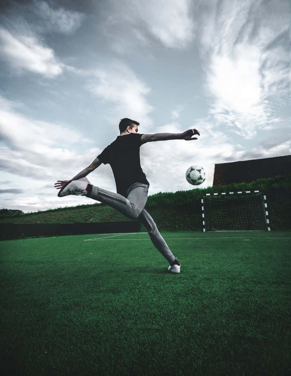3d Sports 桌布 壁紙 — 一名足球運動員在綠色球場上踢球，背景是戲劇性的多雲天空和球門。