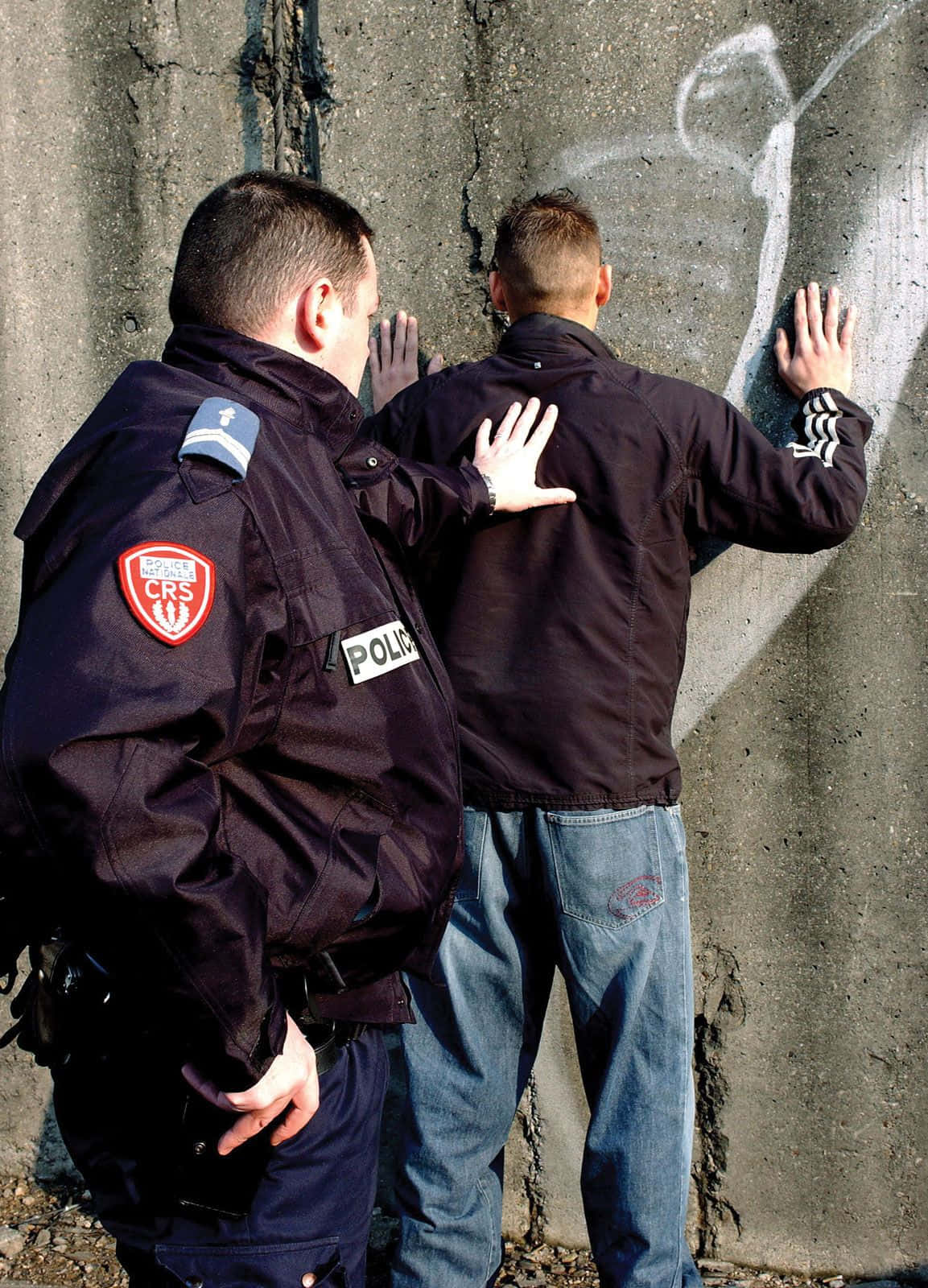 警察 桌布 壁紙 — 一名身穿CRS制服的警員將一名男子按在裂開的混凝土牆上，男子雙手壓在塗鴉覆蓋的表面。