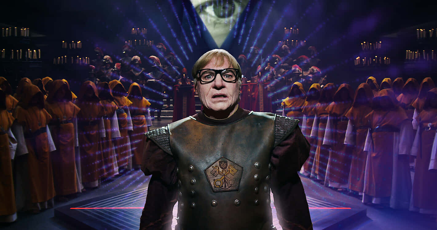 Mike Myers 桌布 壁紙 — 一名身穿中世紀盔甲嘅男人喺紫色戲劇性聚光燈下，企喺舞台中央，前面係一班著住連帽長袍嘅合唱團。
