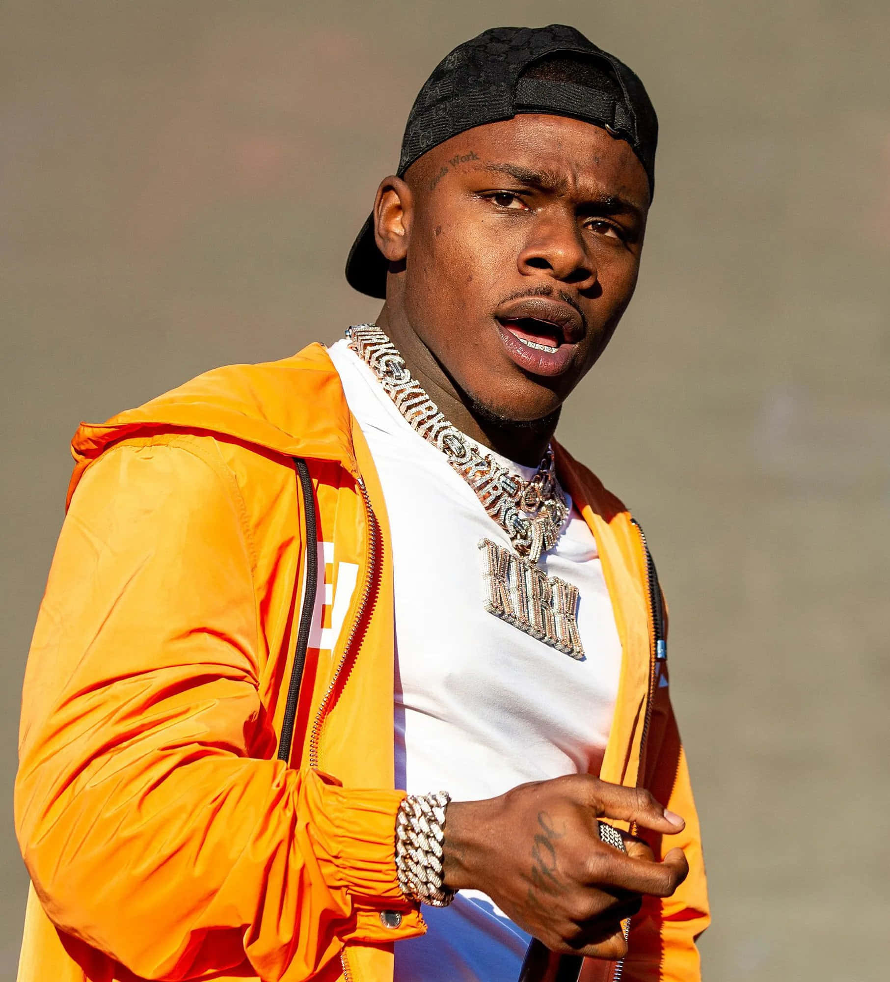 Dababy 桌布 壁紙 — 一名身穿亮橙色外套和黑色帽子的男子，戴著閃亮的鑽石鏈條，專注地看著鏡頭。