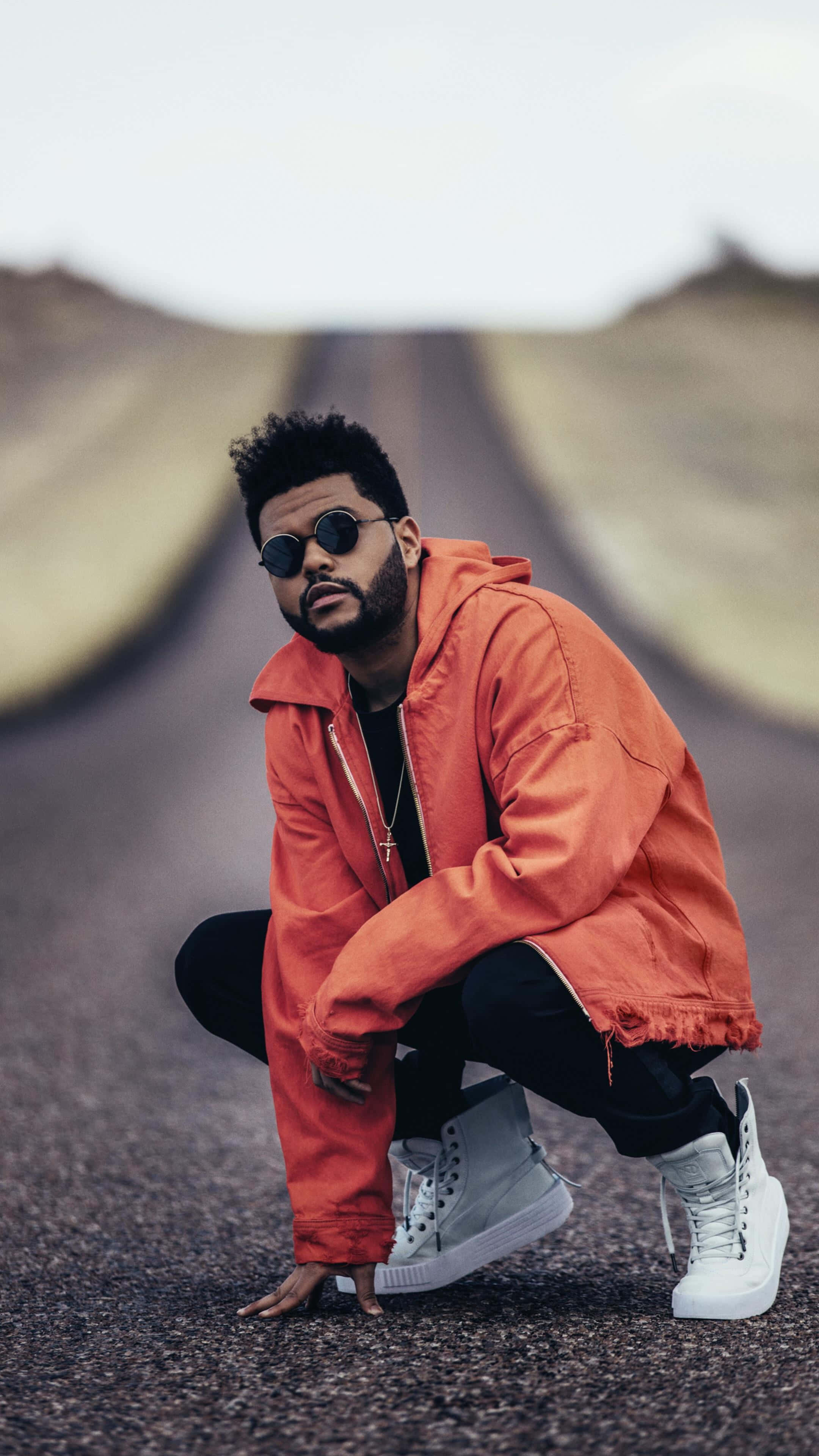 The Weeknd 桌布 壁紙 — 一名身穿亮橙色外套的男子在陰天下的空曠道路上蹲下。