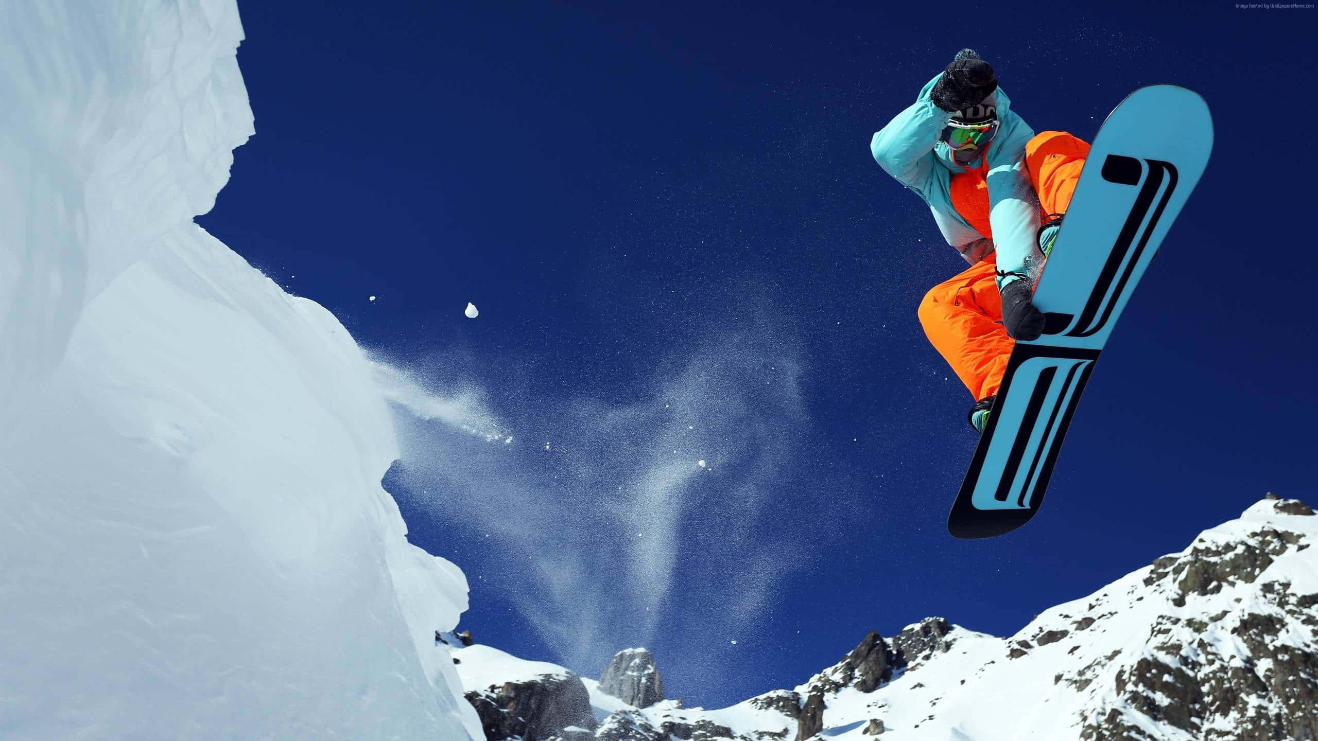Winter Sports 桌布 壁紙 — 一名身穿亮橙色褲和淺藍色外套的滑雪板運動員在白雪覆蓋的山峰上空，在清澈的藍天中翱翔。