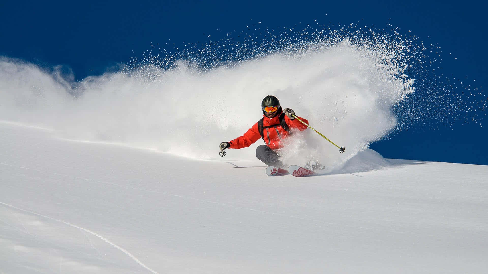 Skiing 桌布 壁紙 — 一名身穿亮紅色外套的滑雪者，在清澈的藍天下，劃過新鮮的粉雪，激起戲劇性的雪霧。