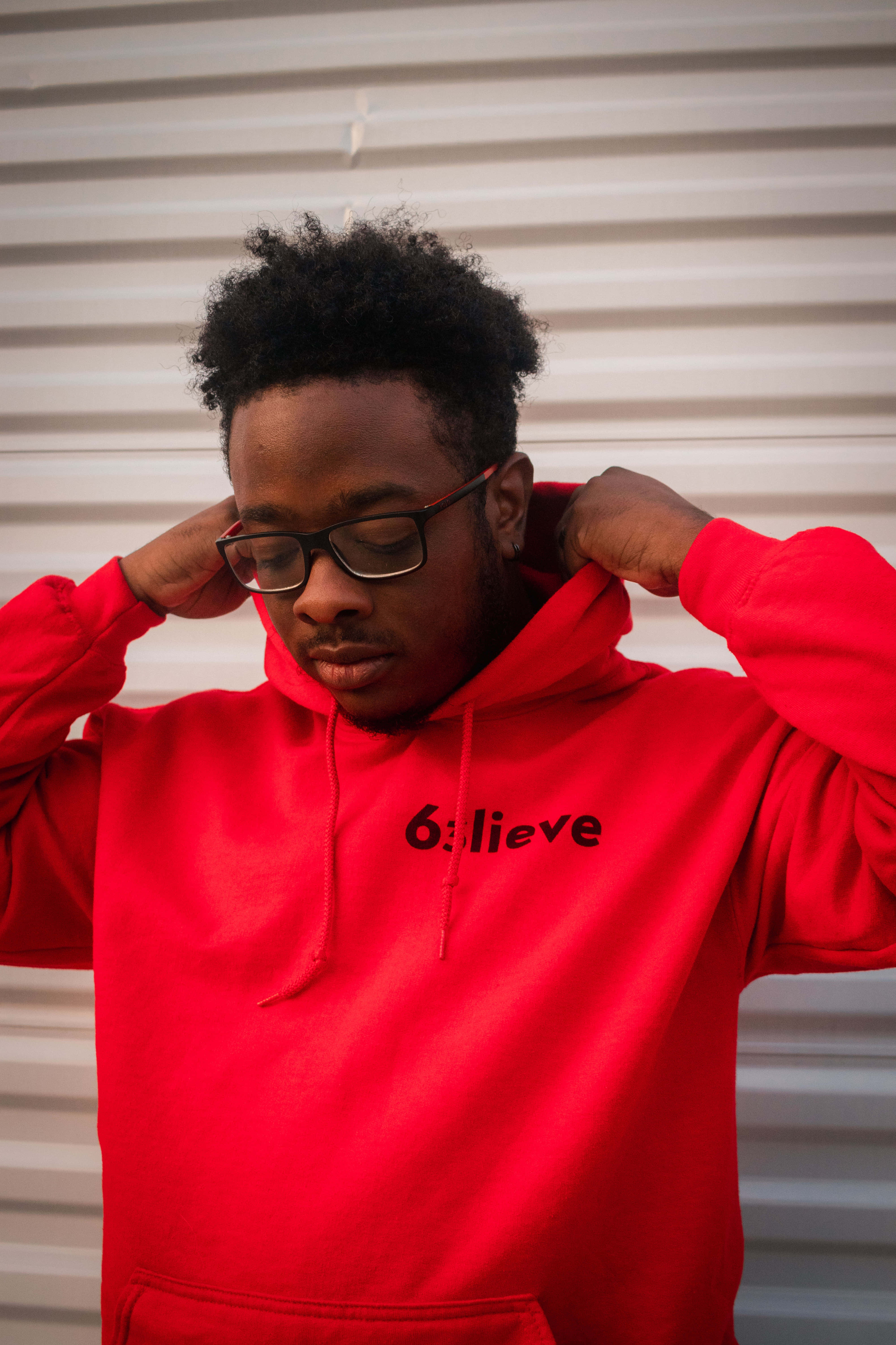Red Hoodie 桌布 壁紙 — 一名身穿亮紅色連帽衫的年輕男子站在白色波紋金屬牆前調整帽子。