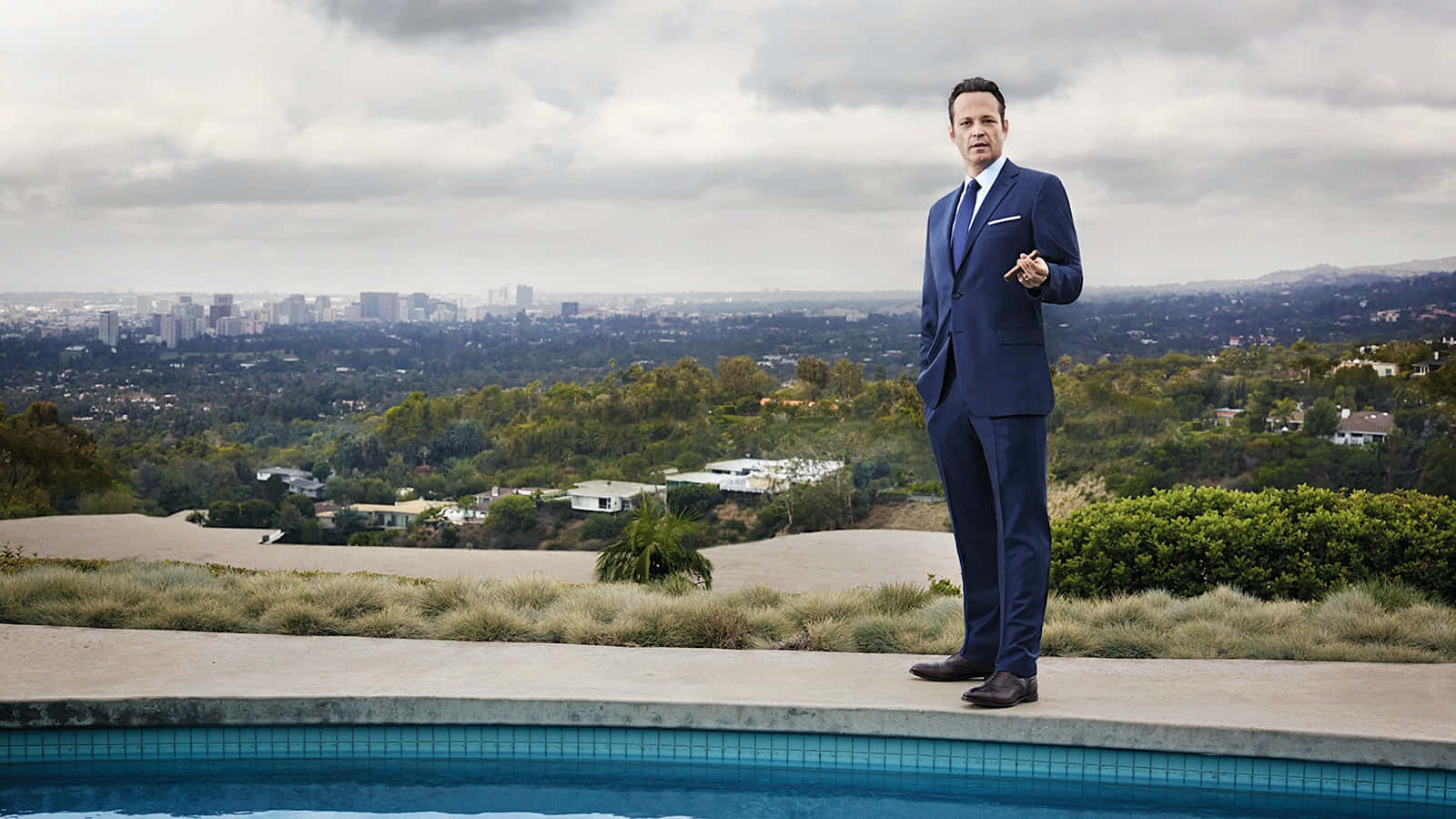 Vince Vaughn 桌布 壁紙 — 一名身穿亮藍色西裝的男子自信地站在無邊際泳池邊緣，俯瞰著陰天下的廣闊城市景觀。