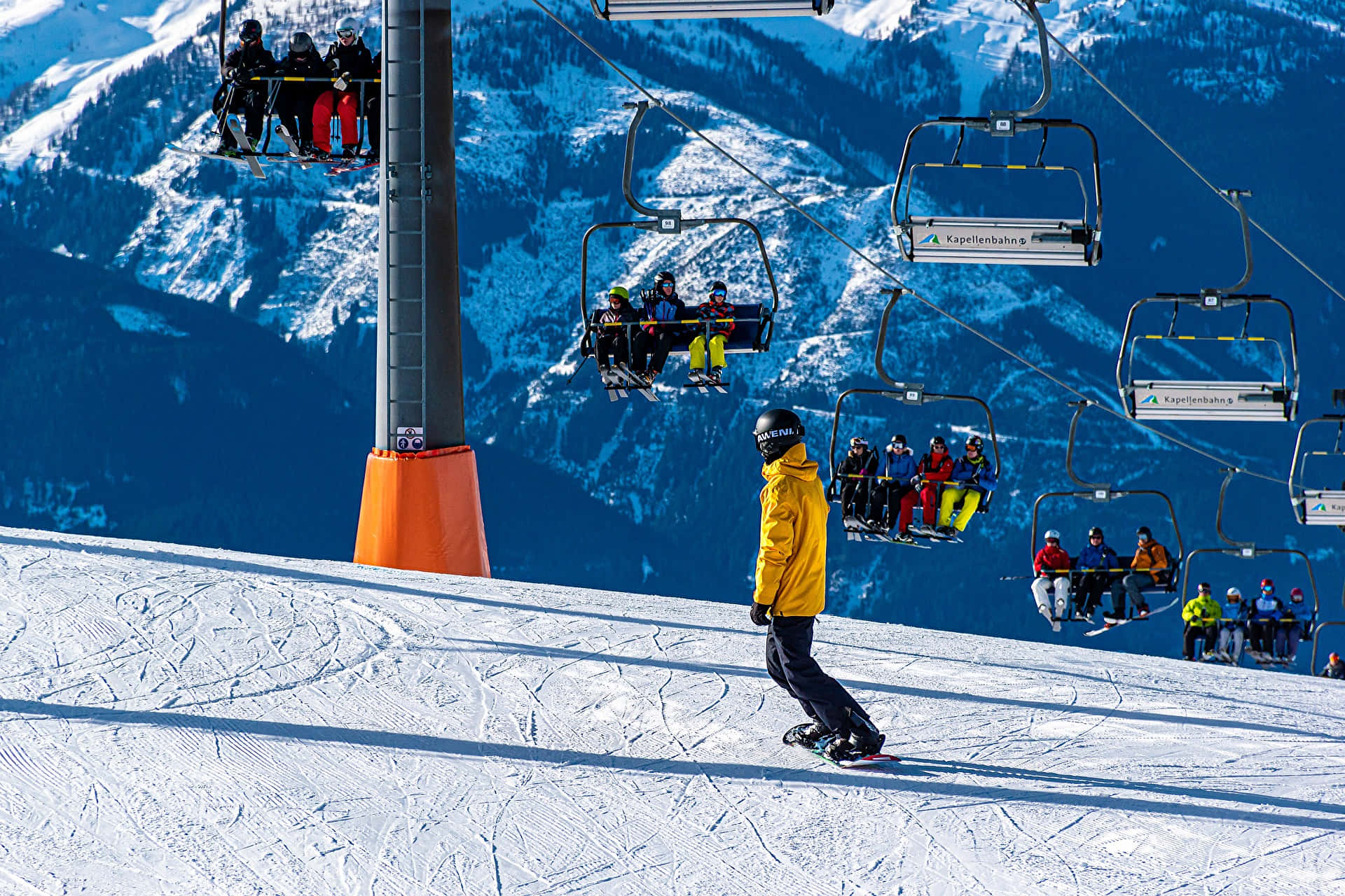 Ski Resort 桌布 壁紙 — 一名身穿亮黃色外套的滑雪板運動員在陽光照耀的斜坡上滑行，背景中纜車載著滑雪者，遠處是白雪覆蓋的山脈。