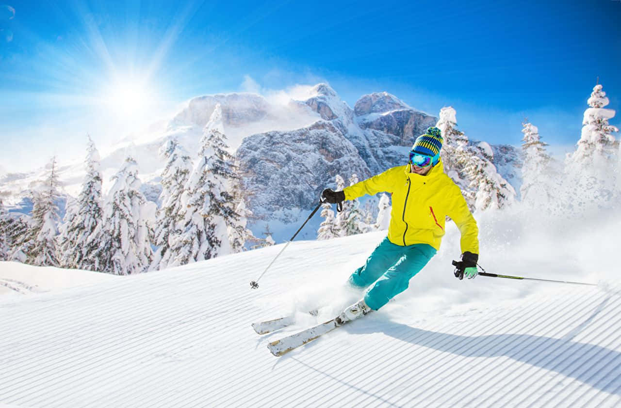 Winter Sports 桌布 壁紙 — 一名身穿亮黃色夾克的滑雪者在陽光明媚的雪坡上穿過新鮮的粉雪，背景是白雪覆蓋的松樹和雄偉的山脈。