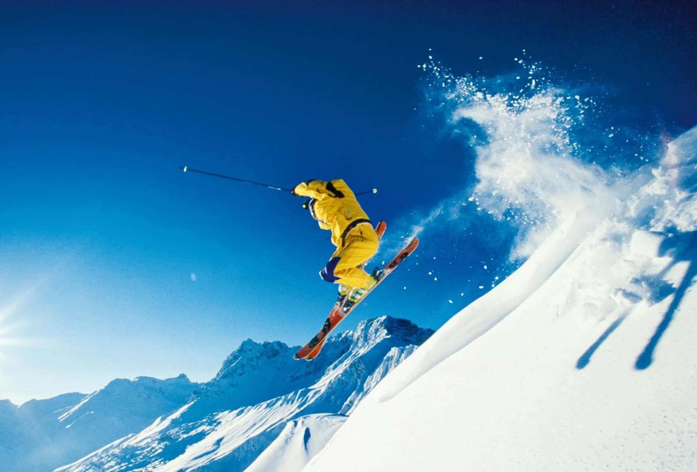 Skiing 桌布 壁紙 — 一名身穿亮黃色滑雪服的滑雪者在晴朗的藍天下，於白雪覆蓋的山坡上空飛翔。