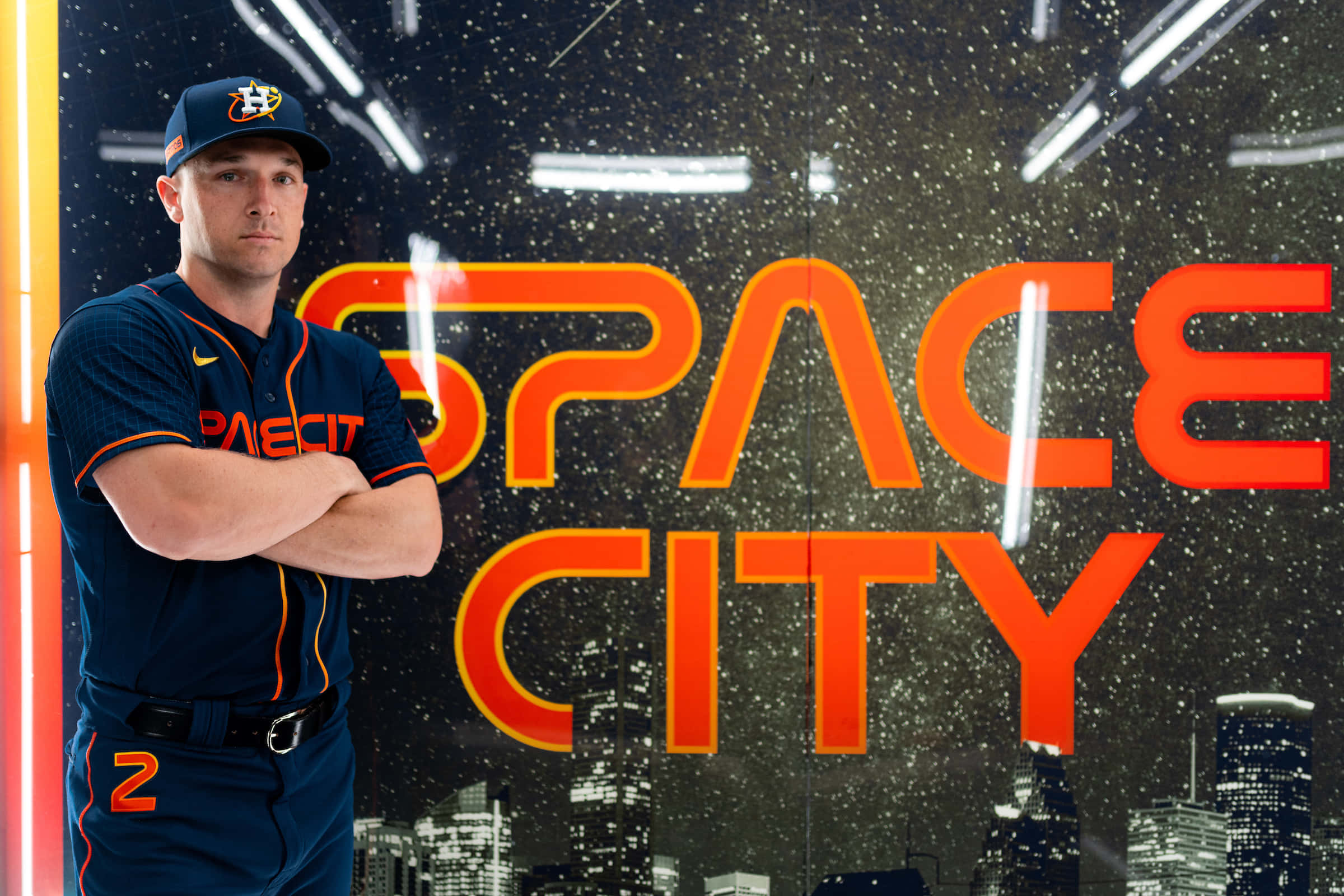Astros 桌布 壁紙 — 一名身穿侯斯頓太空人隊球衣的棒球運動員，在星光璀璨的城市背景下，自信地交叉雙臂站在發光的「SPACE CITY」標誌前。