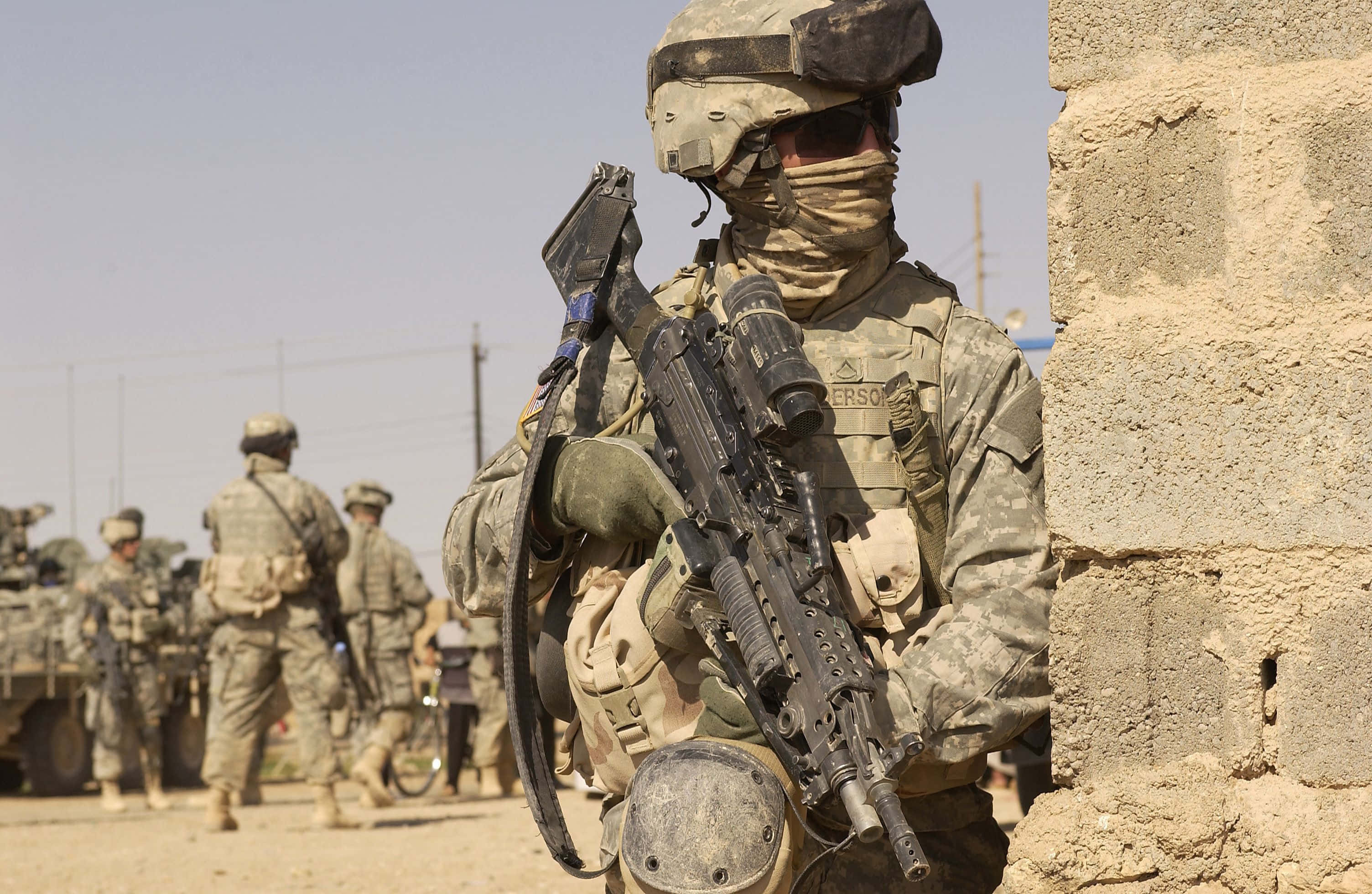 Army 桌布 壁紙 — 一名身穿全副戰鬥裝備的士兵蹲在風化的石牆後面，塵土飛揚的背景中可見其他部隊和裝甲車輛。