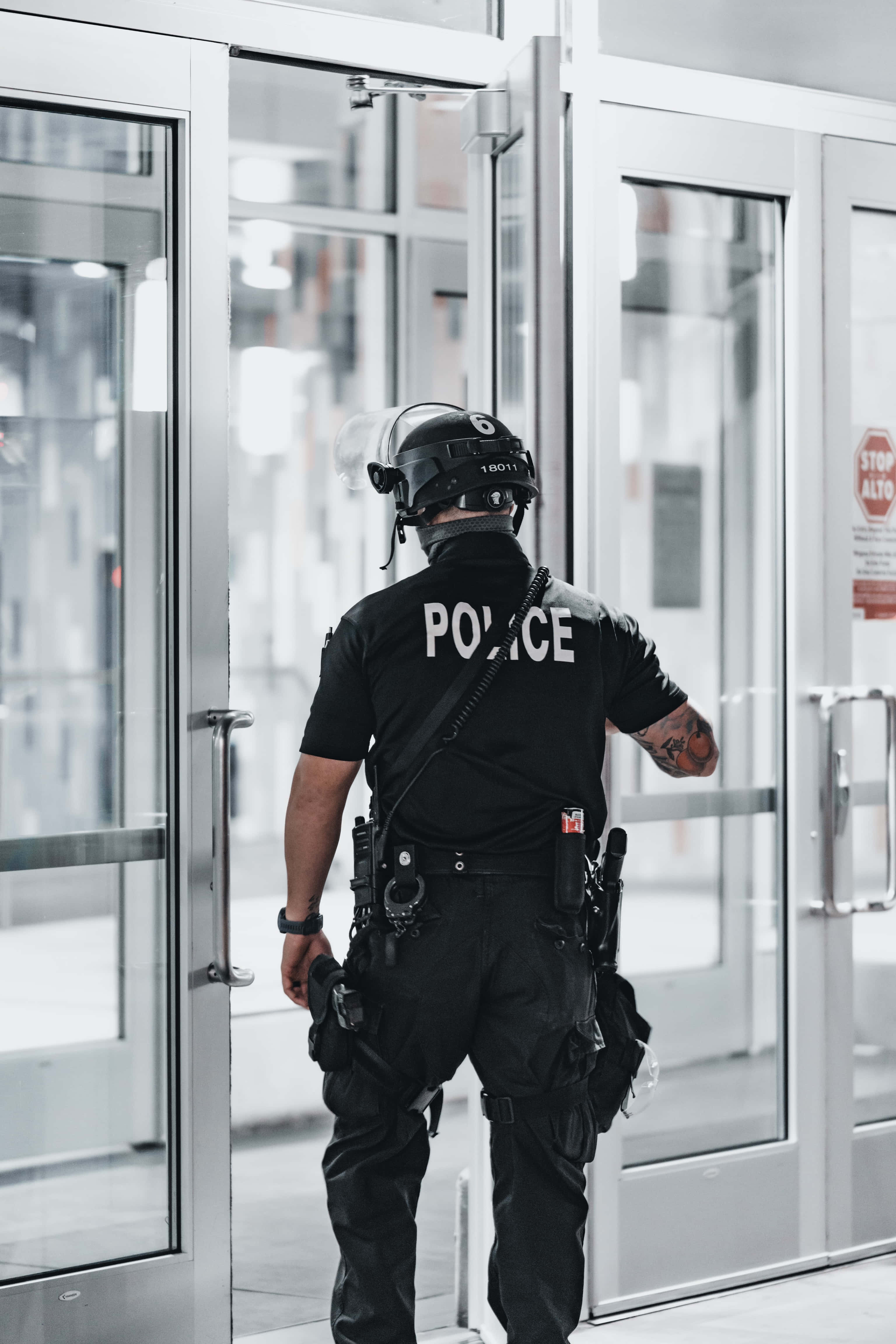 警察 桌布 壁紙 — 一名身穿全套戰術裝備的警察站在現代建築的玻璃門入口處。