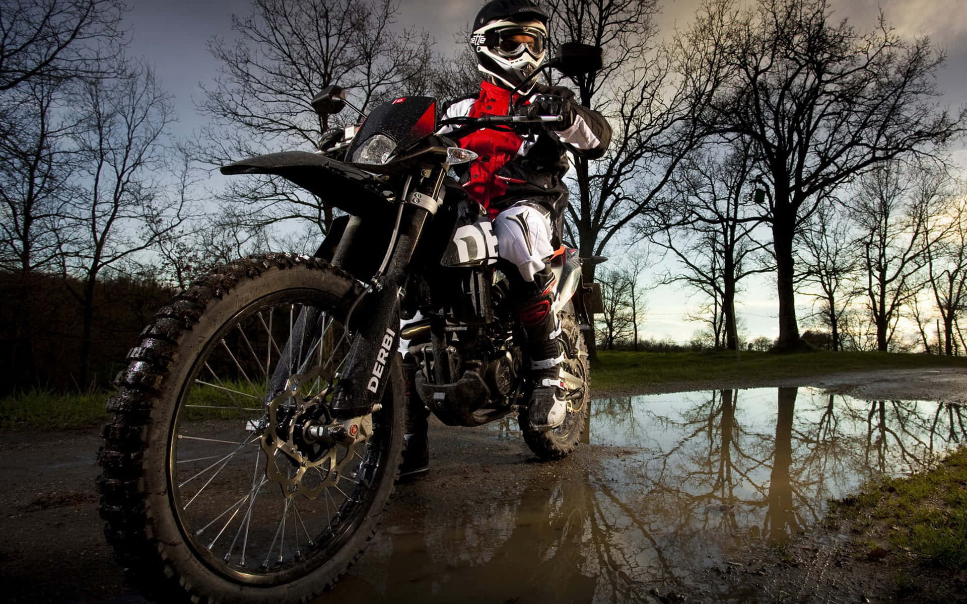 Motocross 桌布 壁紙 — 一名身穿全套裝備的越野電單車手，在陰沉天空下，站在堅固的越野電單車旁，旁邊是一個反射的水坑。