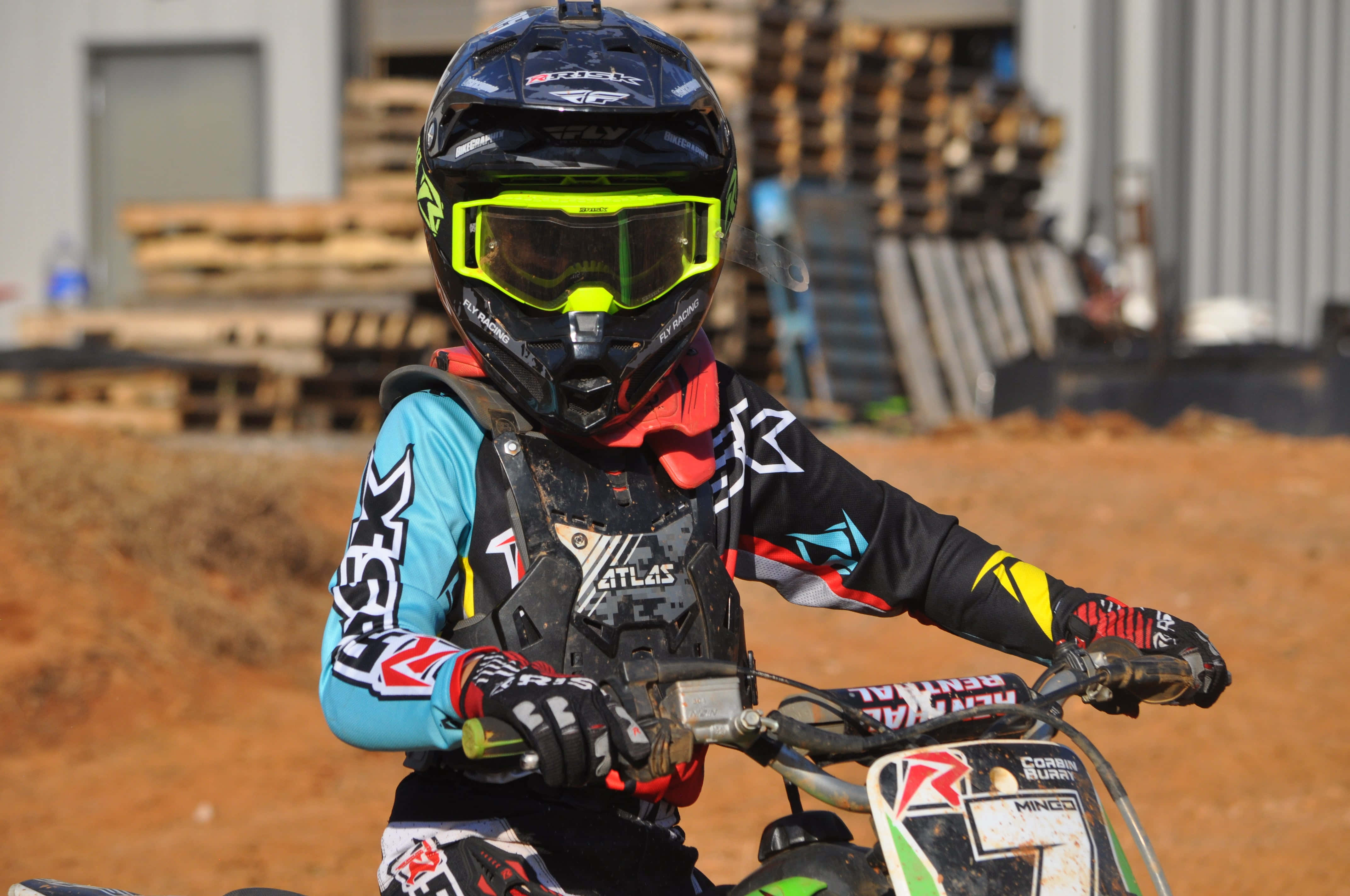 Dirtbike 桌布 壁紙 — 一名身穿全套裝備的越野電單車手在塵土飛揚的賽道上緊握 dirt bike 的把手，背景有堆疊的木製托盤。