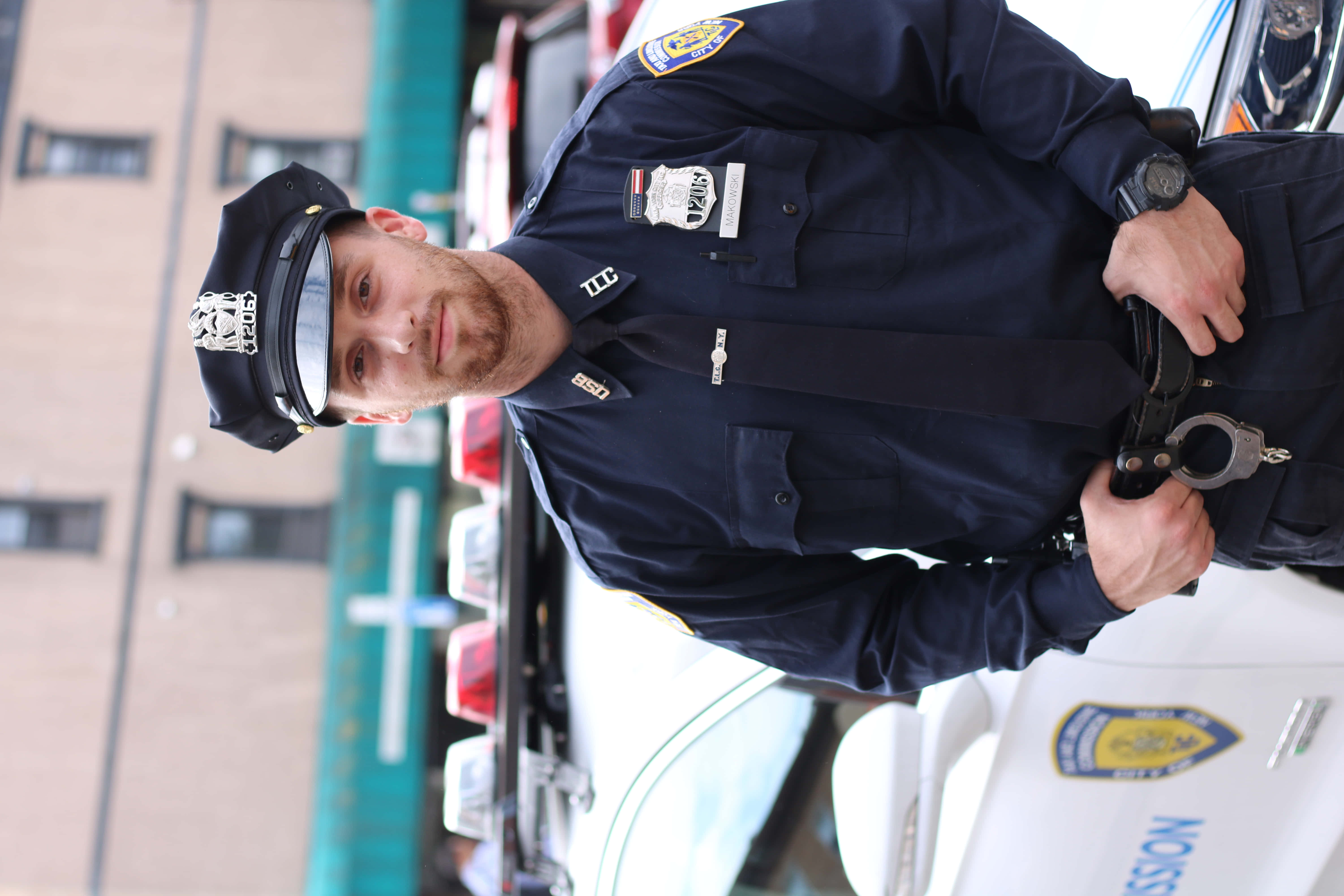 警察 桌布 壁紙 — 一名身穿制服的警察自信地站在巡邏車旁，手持手銬，背景是城市建築。