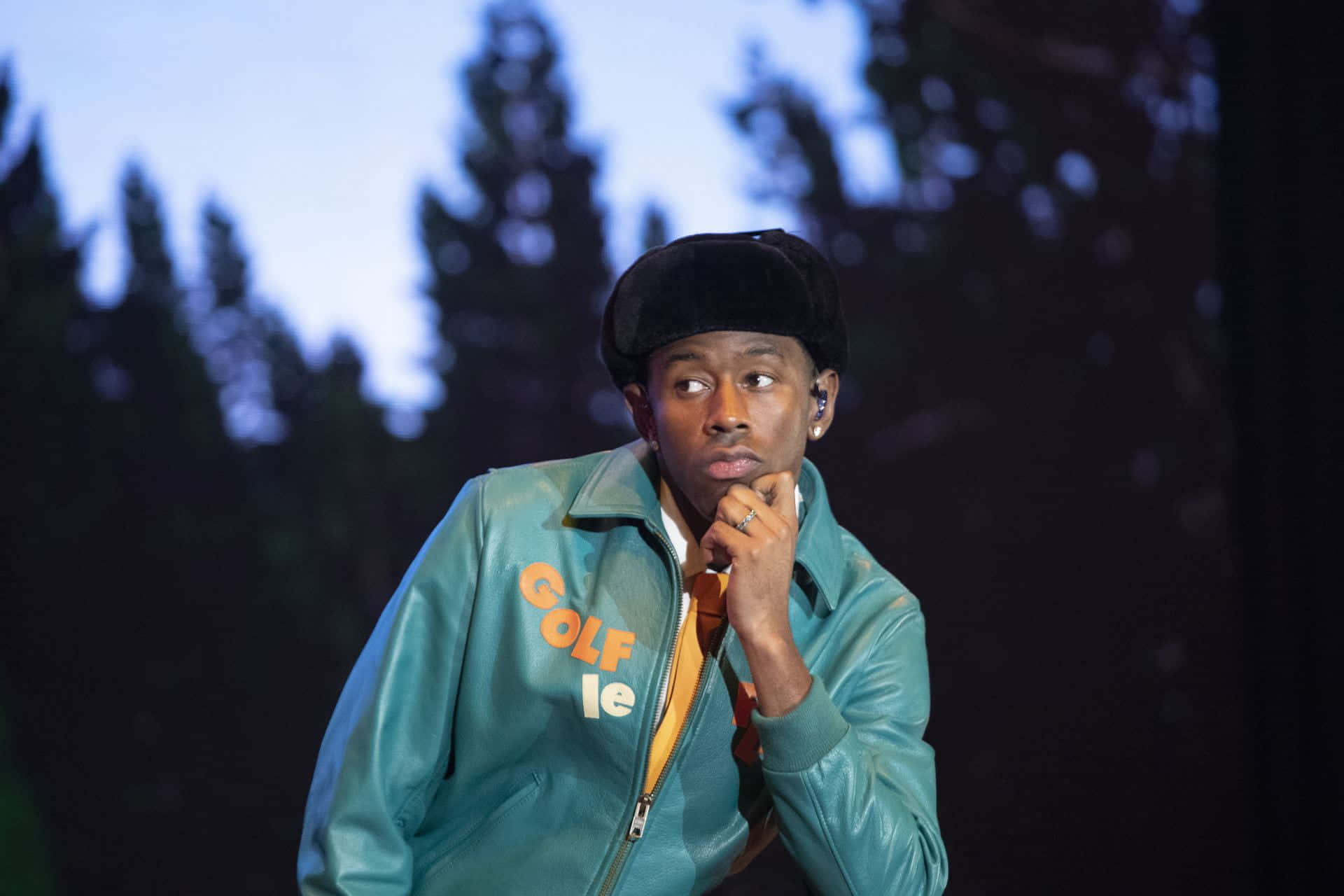 tyler the creator 桌布 壁紙 — 一名身穿印有「GOLF le FLEUR」字樣的青綠色皮褸的男子，在模糊的森林背景下若有所思地向上凝視。