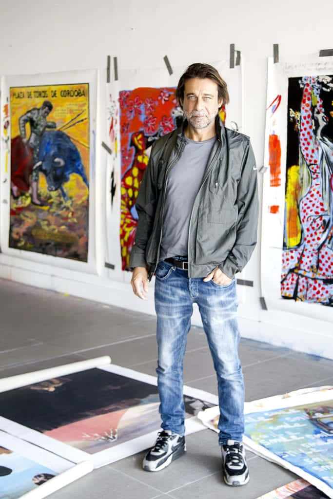 Jordi Mollà 桌布 壁紙 — 一名身穿外套和牛仔褲的男子自信地站在藝術工作室中，周圍牆壁和地板上佈滿了鮮豔的畫作。
