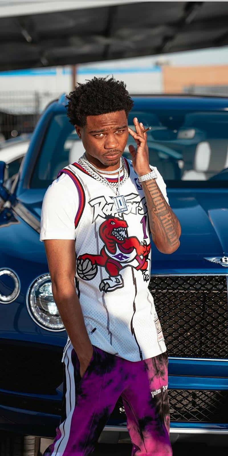 Roddy Ricch 桌布 壁紙 — 一名身穿多倫多速龍隊球衣和紫色紮染褲的年輕男子，自信地在一輛藍色豪華車旁擺姿勢。