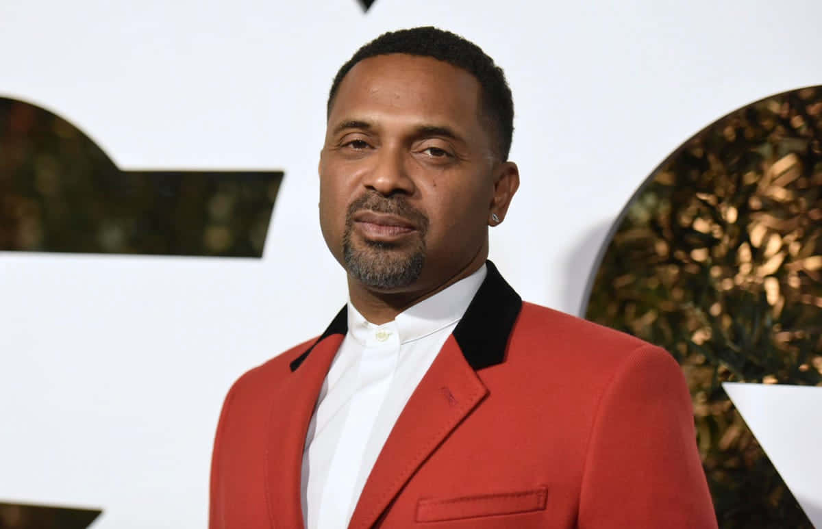 Mike Epps 桌布 壁紙 — 一名身穿大膽紅色西裝、黑色翻領的男子，自信地站在帶有金箔裝飾的白色背景前。