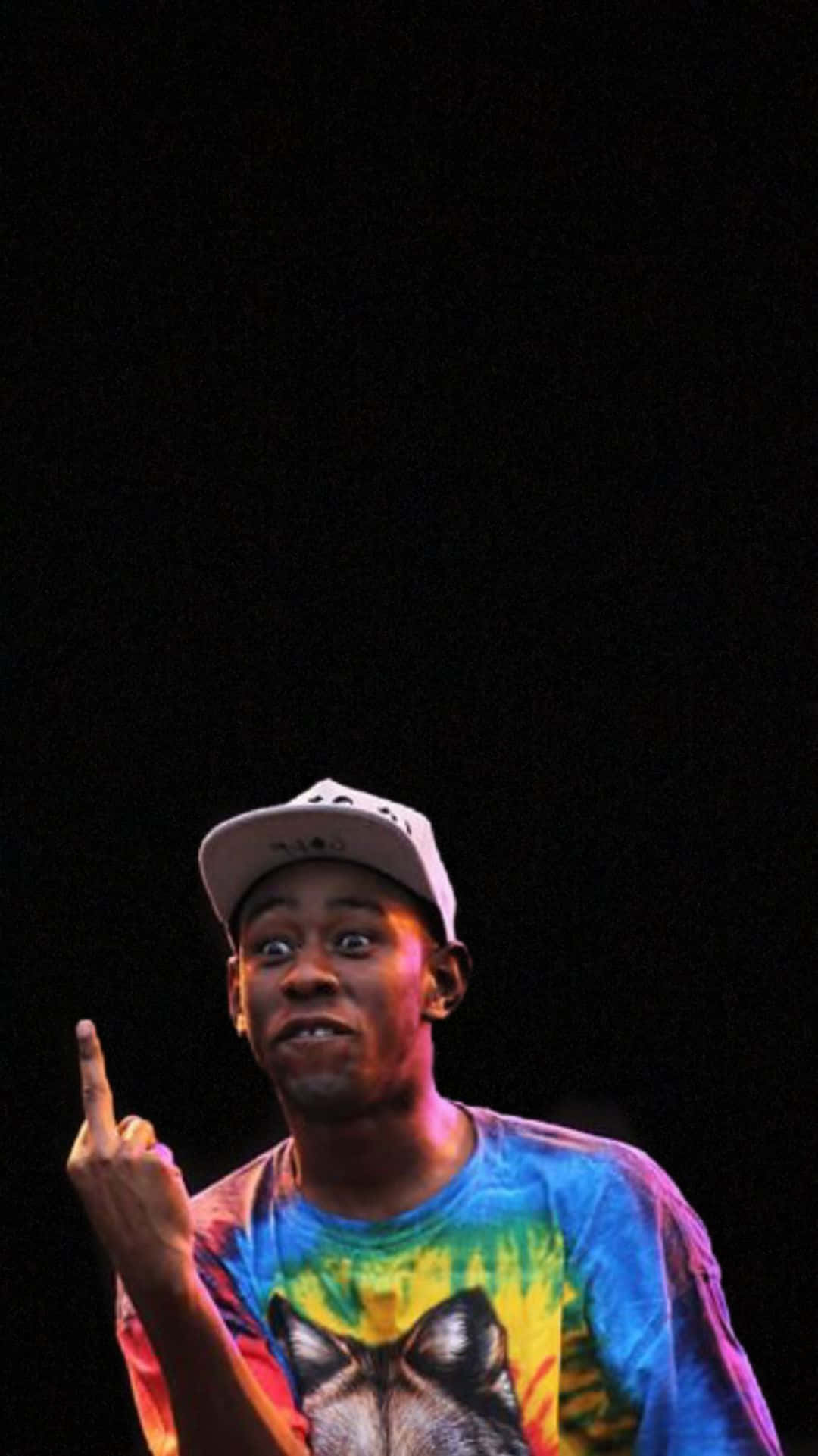 Tyler The Creator - 「When I Say Go」 壁紙 — 一名身穿彩色紮染襯衫和帽子的男子在黑暗背景的舞台燈光下向上指。