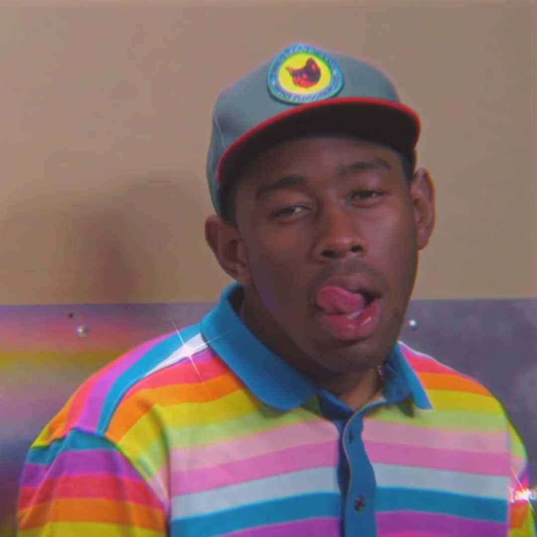 tyler the creator 桌布 壁紙 — 一名身穿彩虹條紋襯衫和帽子的男子，帶著頑皮的表情伸出舌頭。